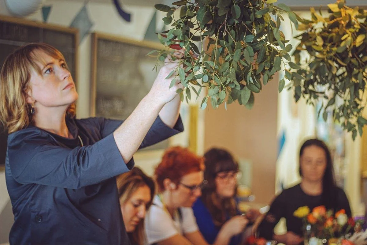 A woman with reddish-brown hair, wearing earrings and a black top, is reaching up to adjust or hang a large leafy green plant or wreath in a room with other women sitting at a table in the background.