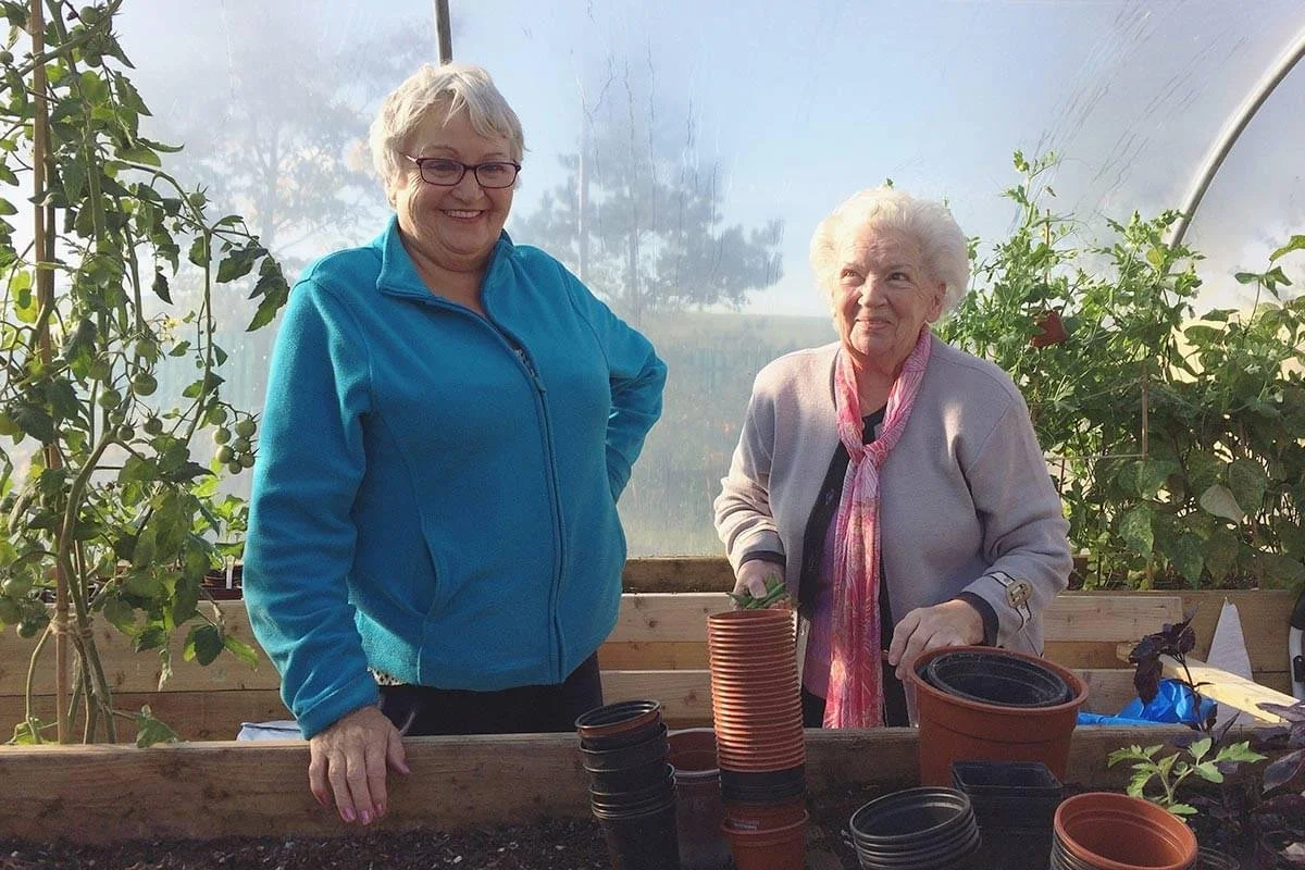 Two women smiling and working with potted plants inside a greenhouse, surrounded by gardening supplies and plants.
