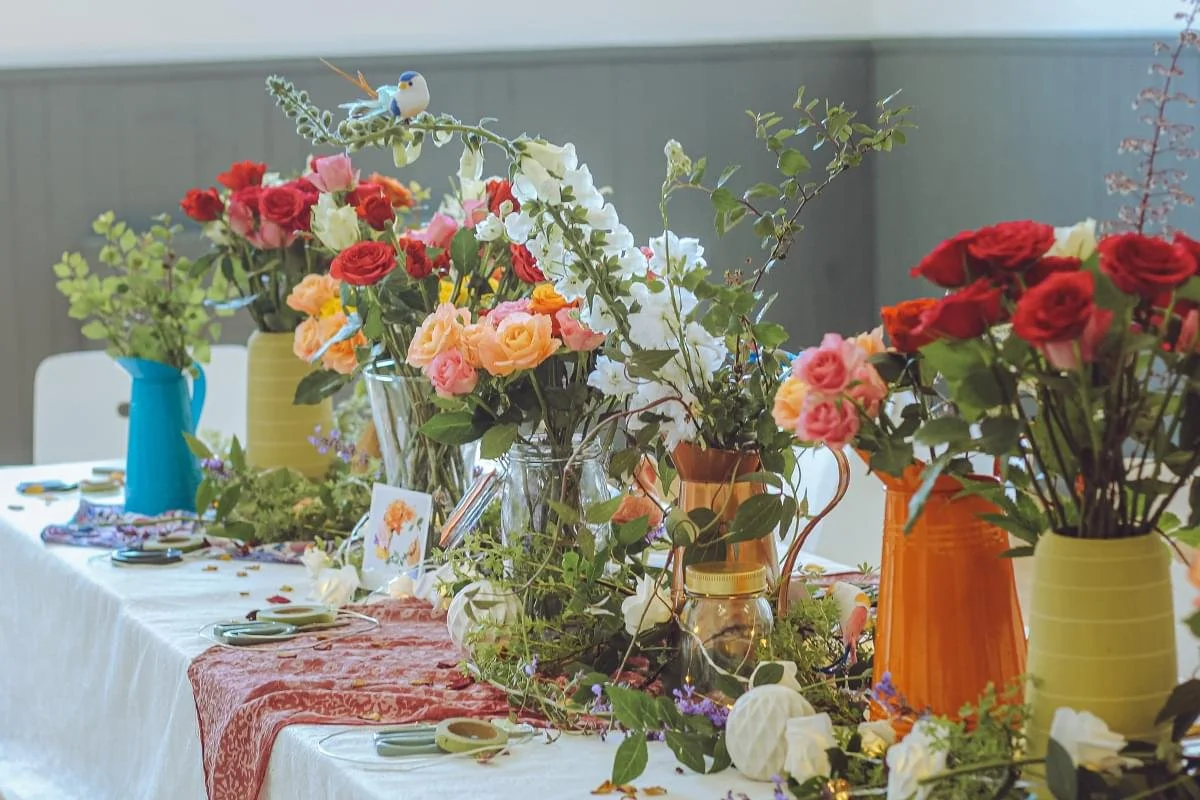 Table decorated with colorful vases and flower arrangements, including roses and greenery, set up for a celebration or event.