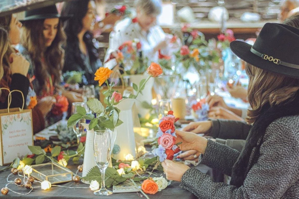 Group of women at a table decorated with flowers, candles, and fairy lights, engaging in craft activities, with some wearing hats.