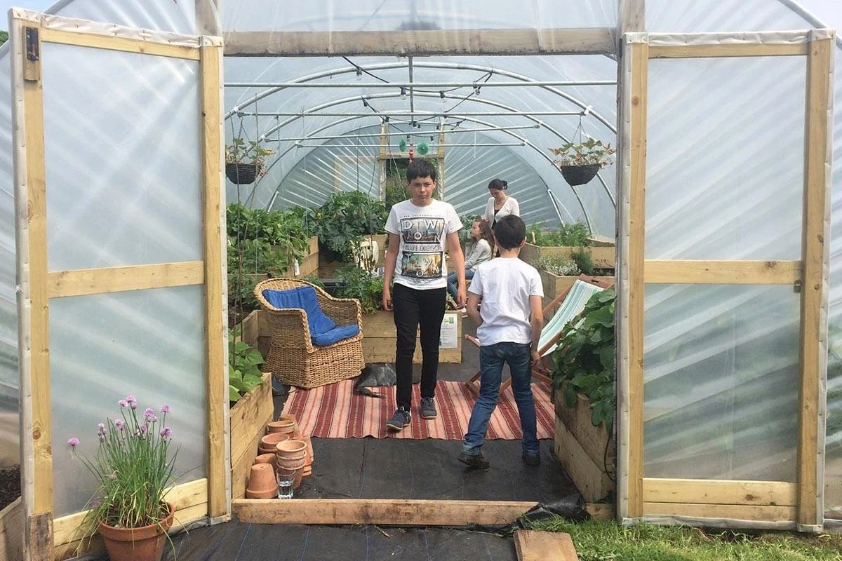People inside a greenhouse with plants and gardening supplies, including flower pots and hanging baskets, and a wicker chair with a blue cushion.