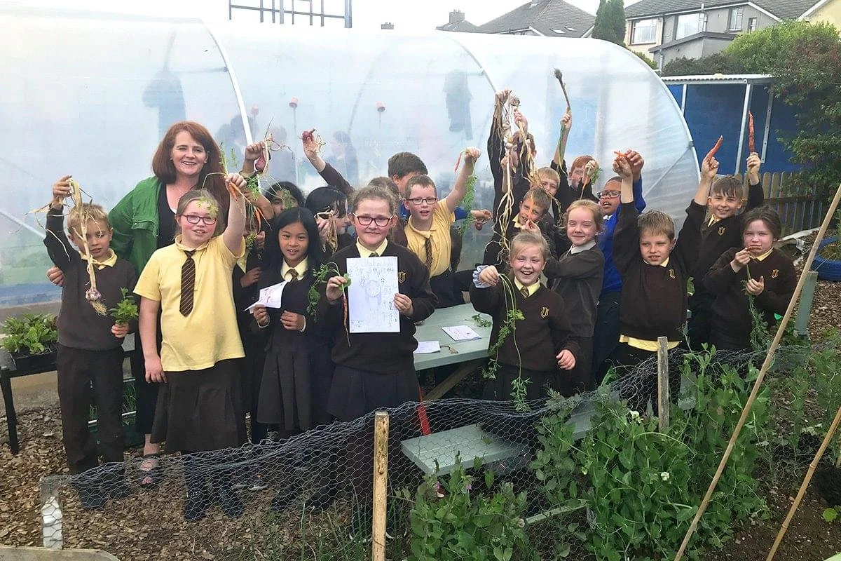 Group of children and a woman in a garden, holding up freshly harvested vegetables and smiling, with a greenhouse behind them.