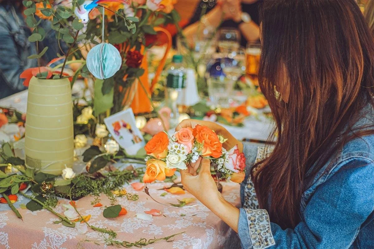 A woman with long brown hair, wearing a denim jacket, is arranging orange, white, and peach-colored flowers into a bouquet at a decorated table with flowers, candles, and decorative items.