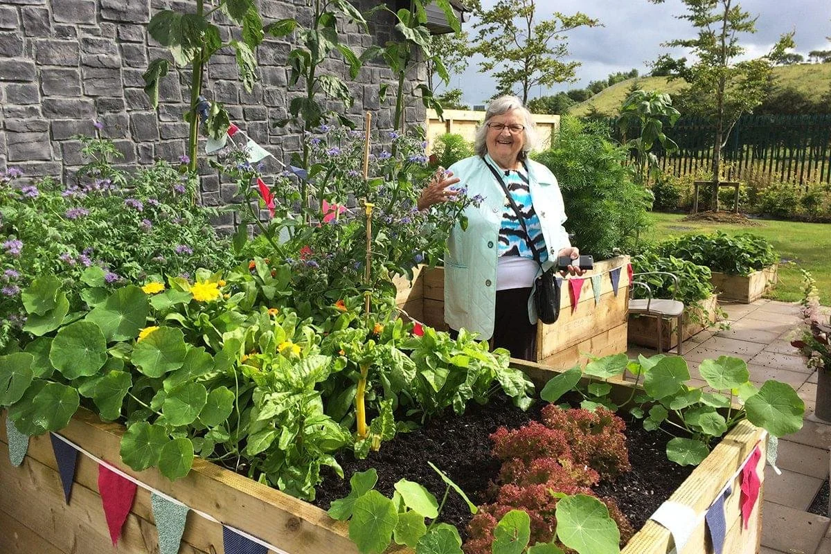 An elderly woman smiling in her garden, surrounded by lush green plants and colorful flowers, standing next to wooden planters filled with soil and plants, with a stone wall and trees in the background.