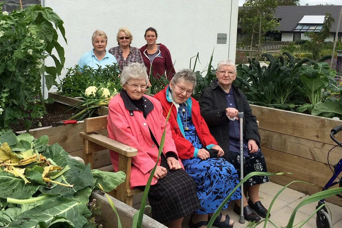 Four elderly women sitting on a wooden bench in a garden, surrounded by leafy plants and vegetables. Two women are standing behind them, posing for the photo.