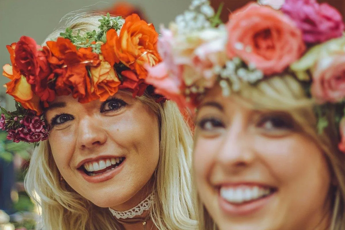 Two women wearing flower crowns made of colorful roses and other flowers, smiling and smiling at the camera.