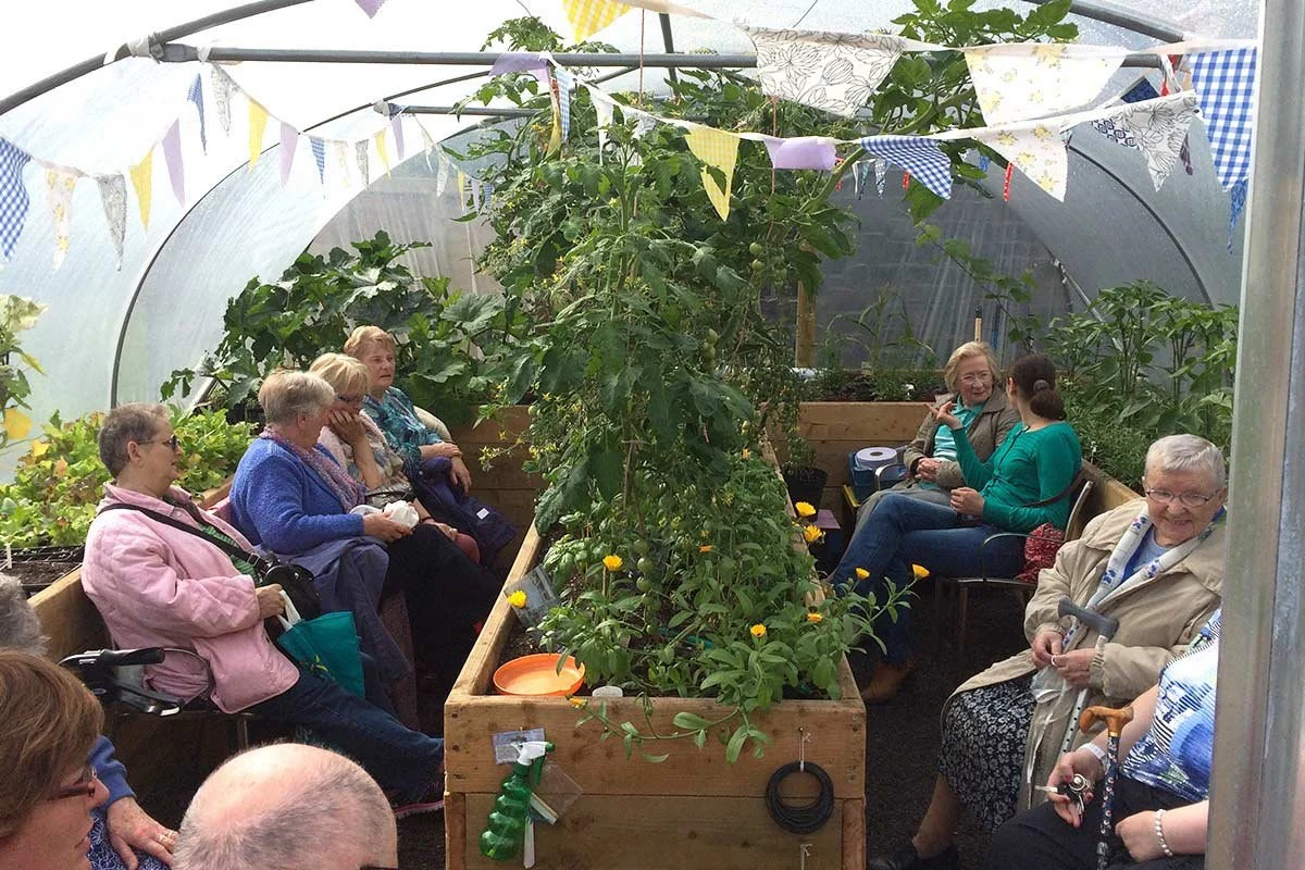 Group of elderly women sitting inside a greenhouse surrounded by plants, some engaged in conversation, others sitting quietly, decorated with colorful bunting and banners hanging from the ceiling.