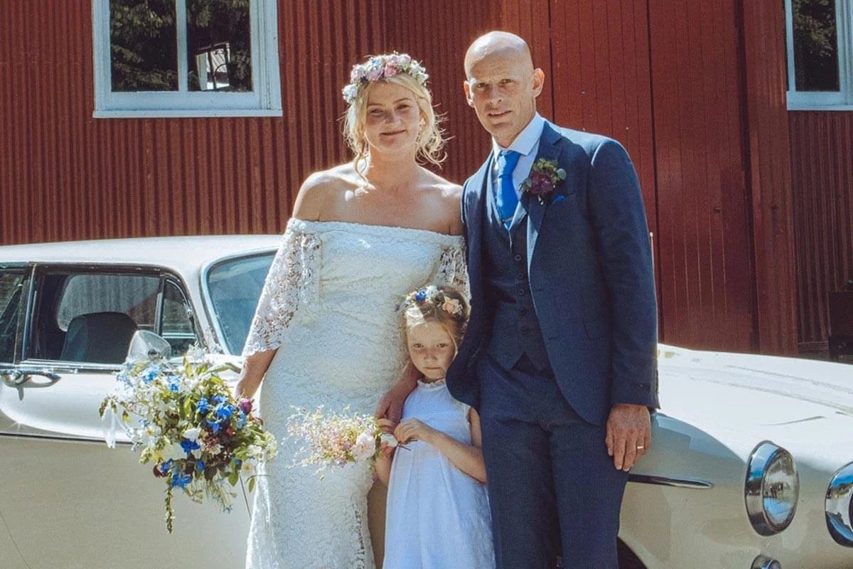 A bride, groom, and young girl at a wedding in front of a vintage white car and a red wooden building, with the bride holding a bouquet of flowers and the young girl holding a small floral arrangement.