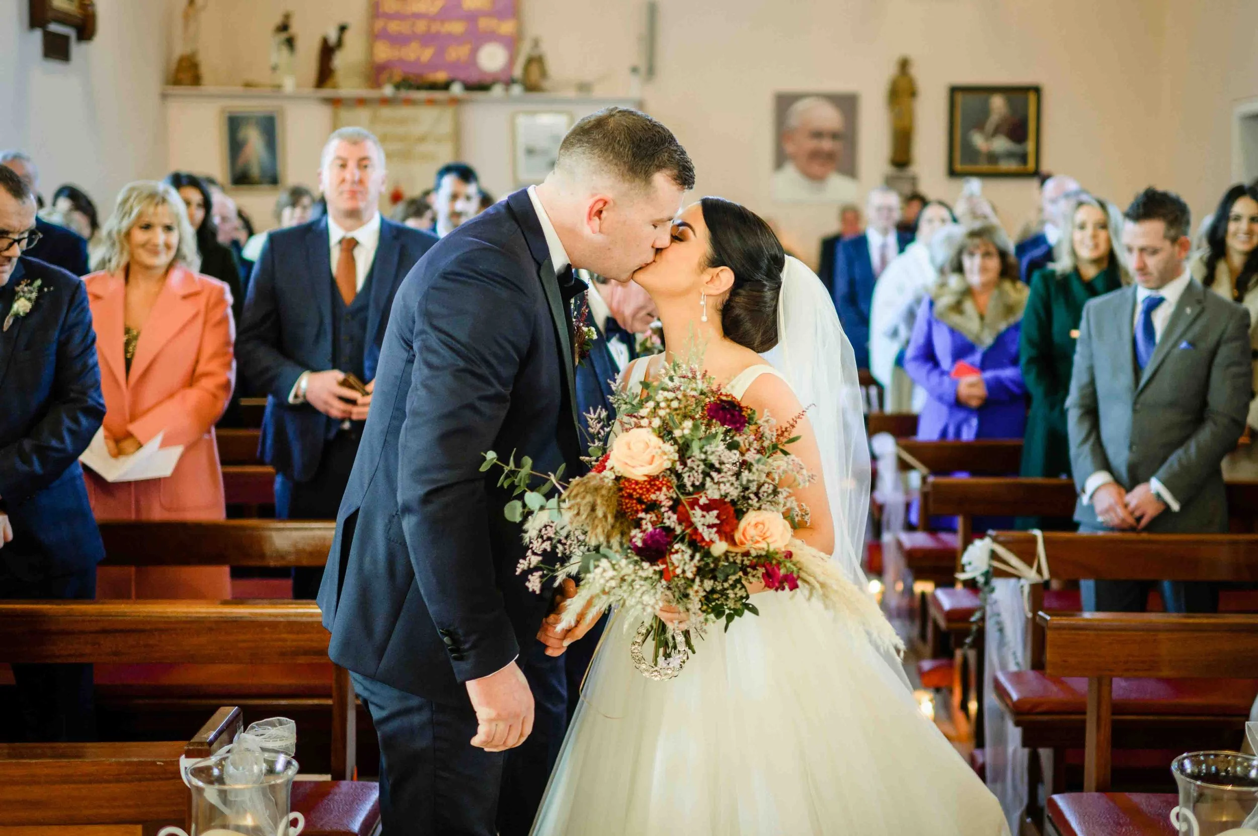 A bride and groom share a kiss in a church during their wedding ceremony, surrounded by seated guests.