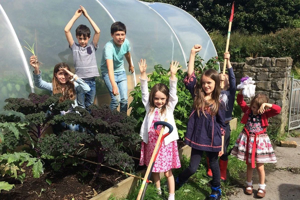 Children gardening outside near a greenhouse, showing excitement and happiness.