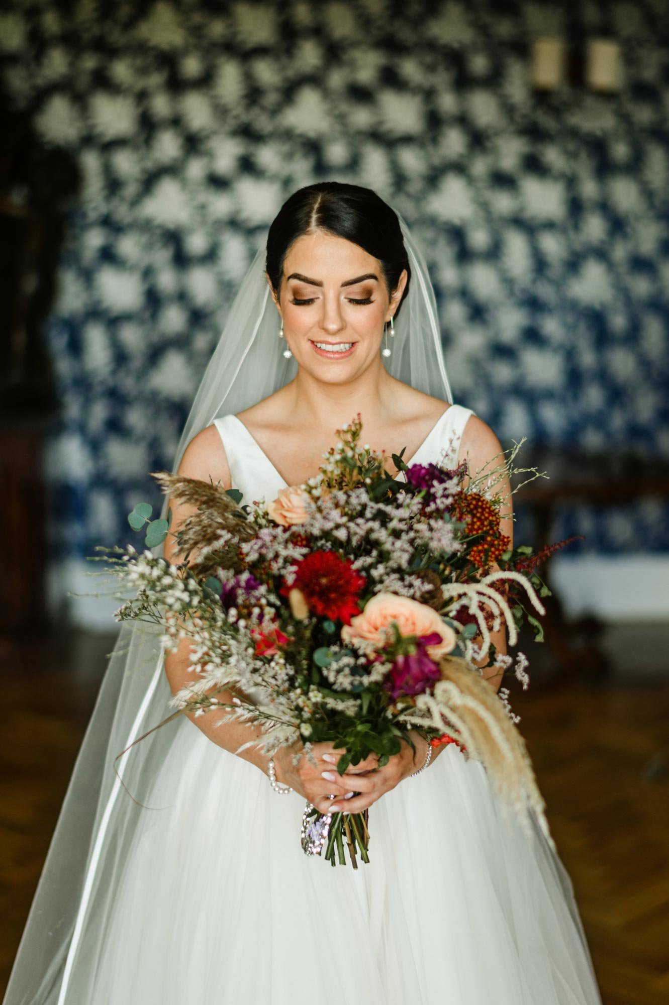 A smiling bride holds a large colorful bouquet of flowers, wearing a white wedding dress and veil, with a dark patterned wall background.