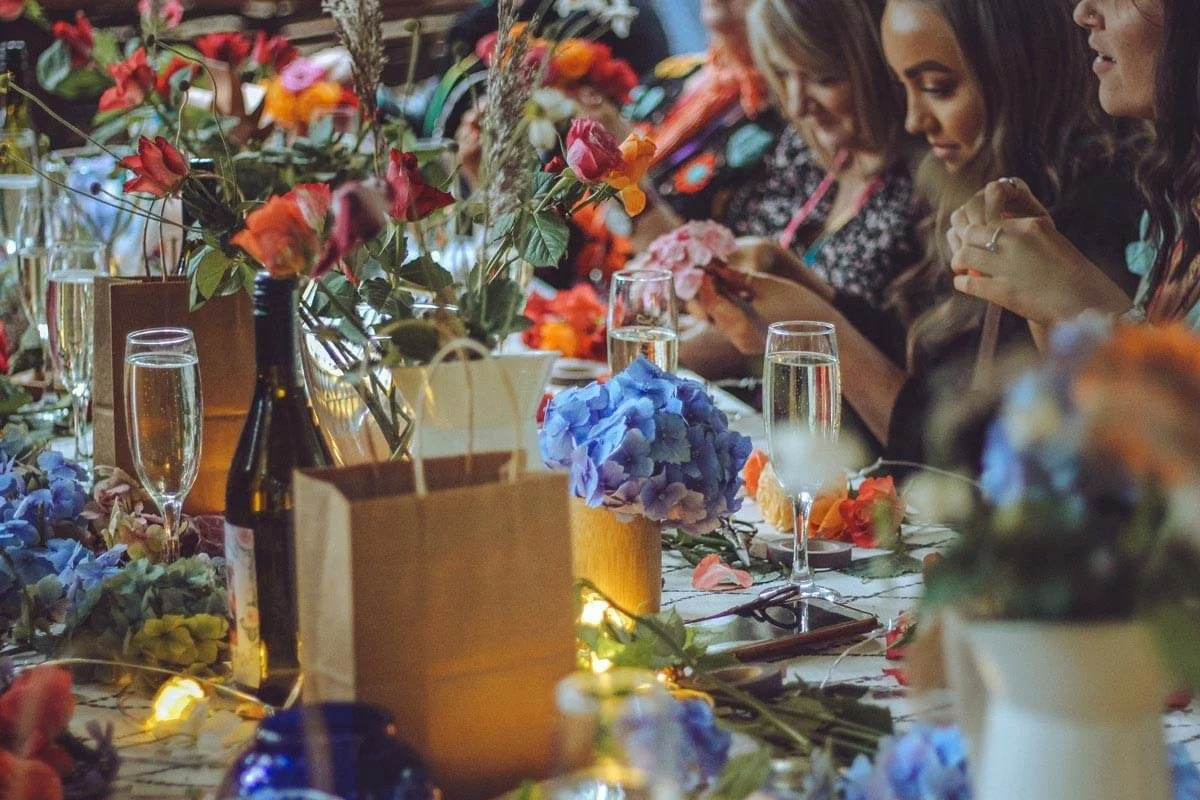 People sitting at a table decorated with colorful flowers and vases, enjoying a celebration with glasses of champagne.
