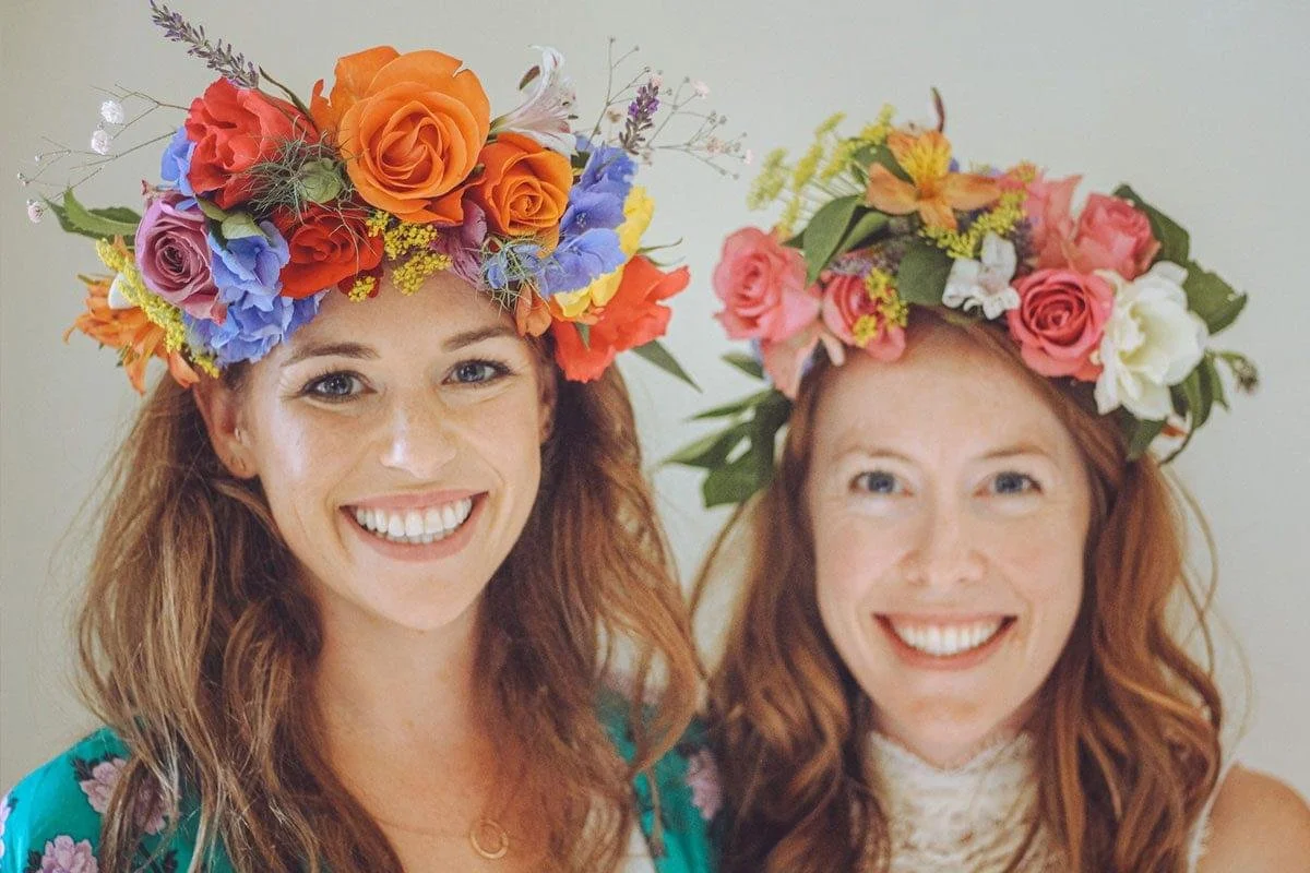 Two women smiling with flower crowns, both with reddish hair, one wearing a teal floral dress and the other wearing a white lace top.