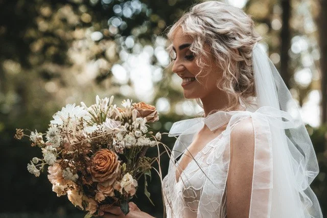 Bride with curly blonde hair holding a bouquet of flowers outdoors.