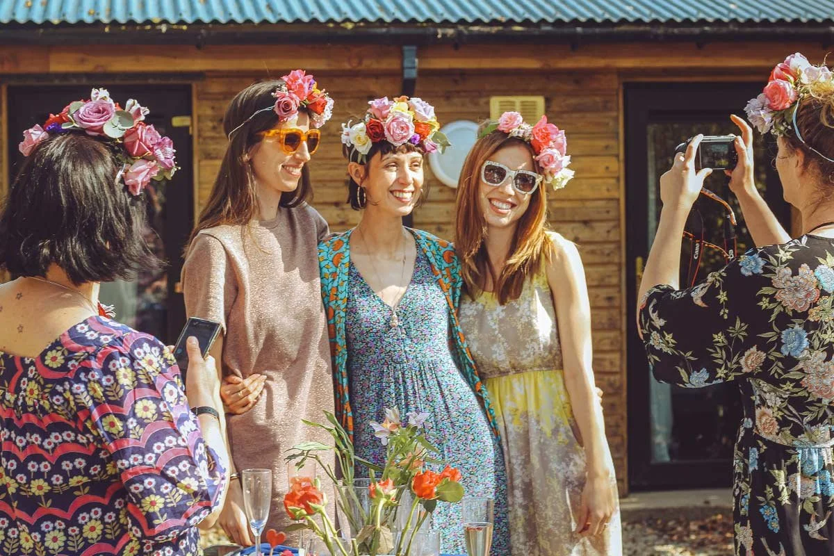 Four women wearing flower crowns pose for a photo during a celebration, outdoors with a wooden building in the background.