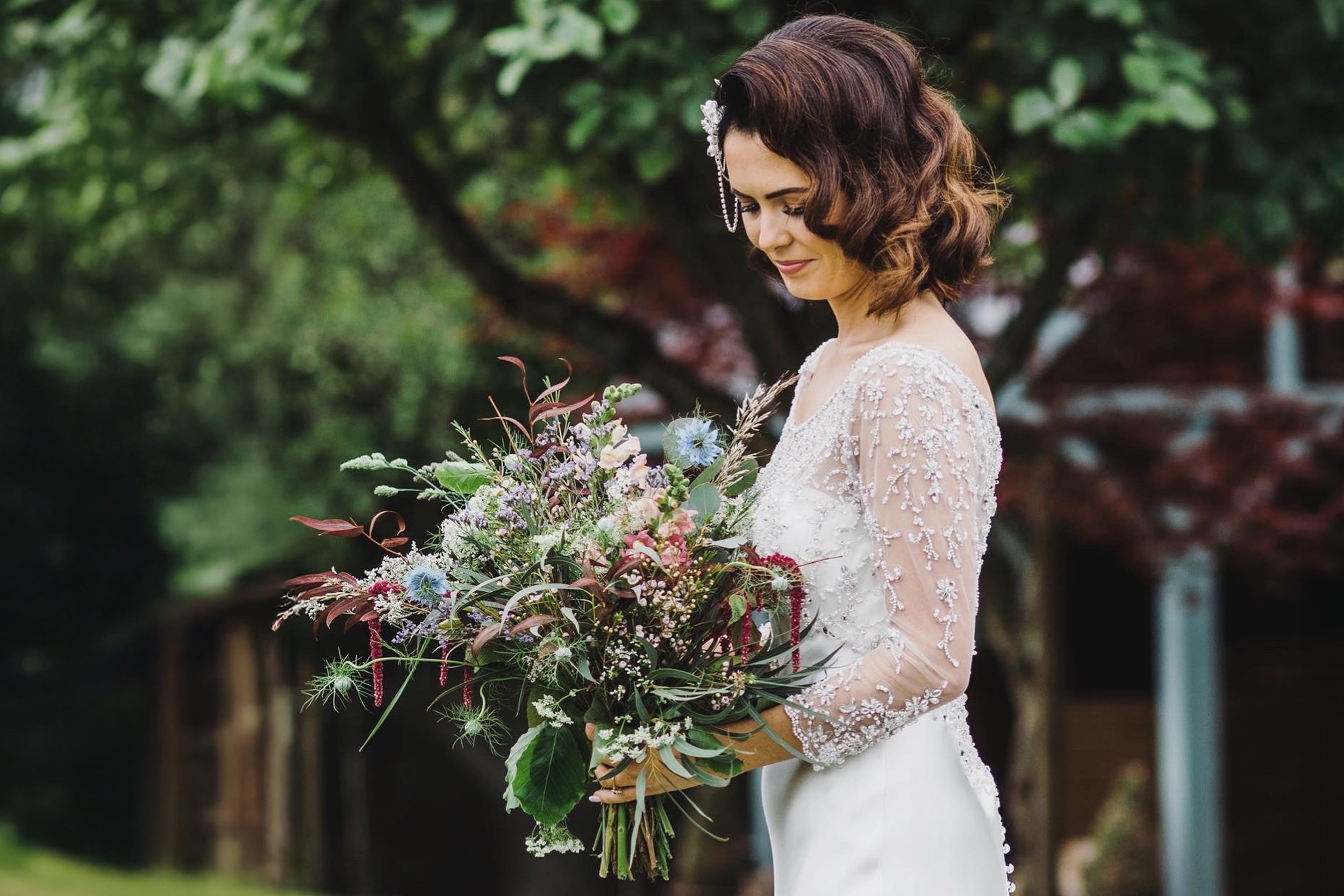 A bride with shoulder-length brown hair and a floral hairpiece, wearing a white wedding dress with lace and beaded embellishments on the sleeves and bodice, holding a large bouquet of pink, purple, white, and blue flowers, standing outdoors in a gard