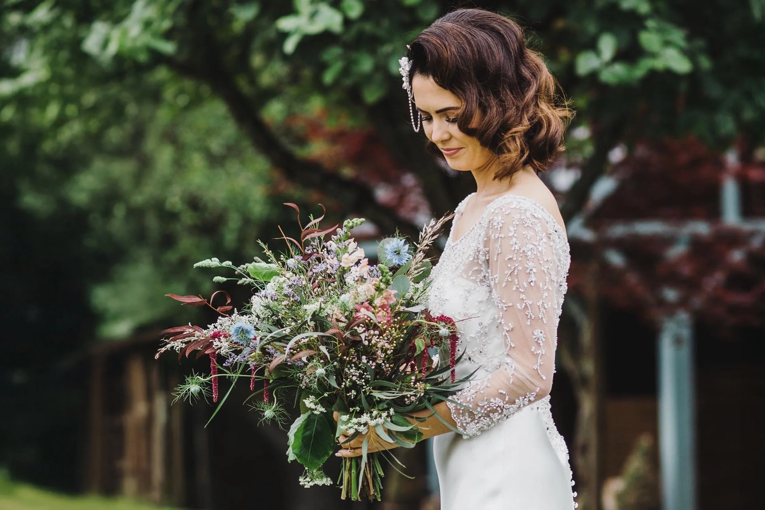 A woman in a white wedding dress with lace sleeves holding a large colorful bouquet of flowers outdoors in a garden with green trees in the background.
