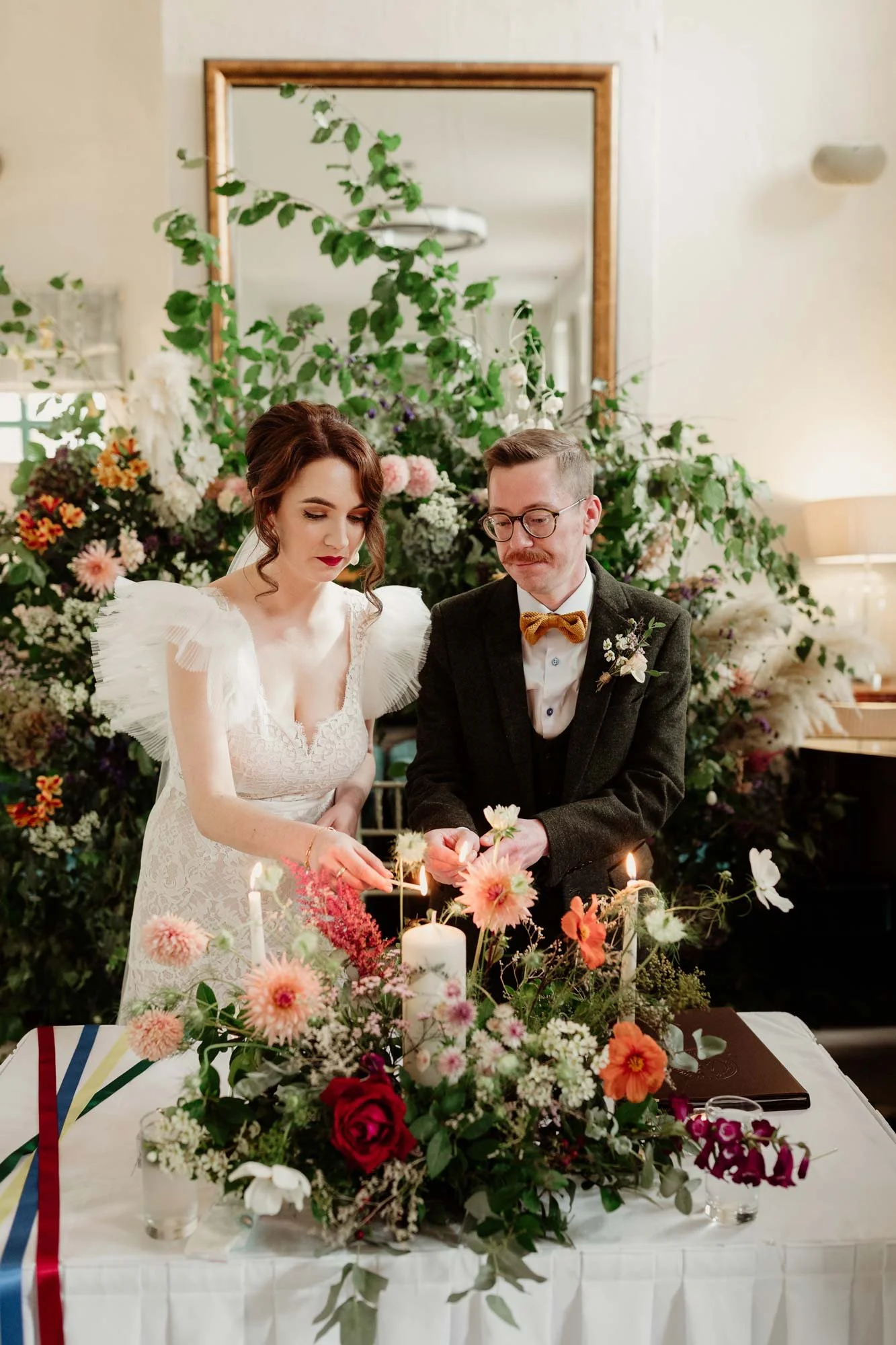 A bride and groom lighting candles during their wedding ceremony, surrounded by a large floral arrangement on a table.