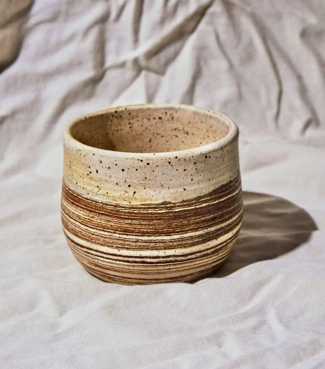 A round, handmade ceramic cup with horizontal brown and beige stripes, placed on a white fabric background with crumpled white paper behind it.