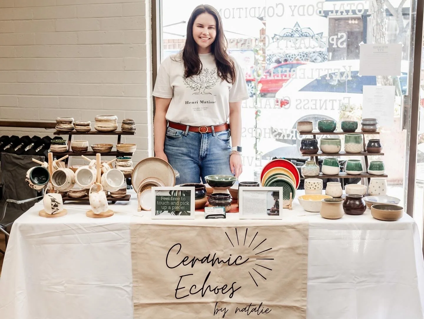 A woman standing behind a table with various ceramic bowls and pottery on display at a ceramic jewelry booth called 'Ceramic Echoes by natalie'.