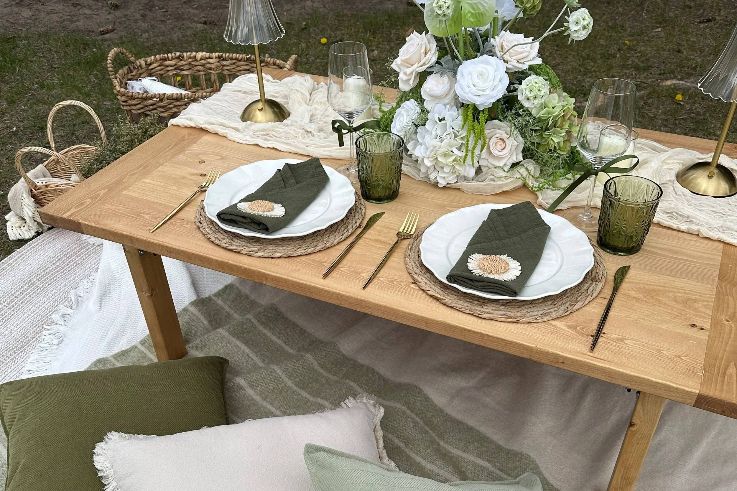 An outdoor luxury picnic set up with a natural, organic theme featuring greens and whites. Includes green drinking glasses, wine glasses, candles, and brass lamps .
