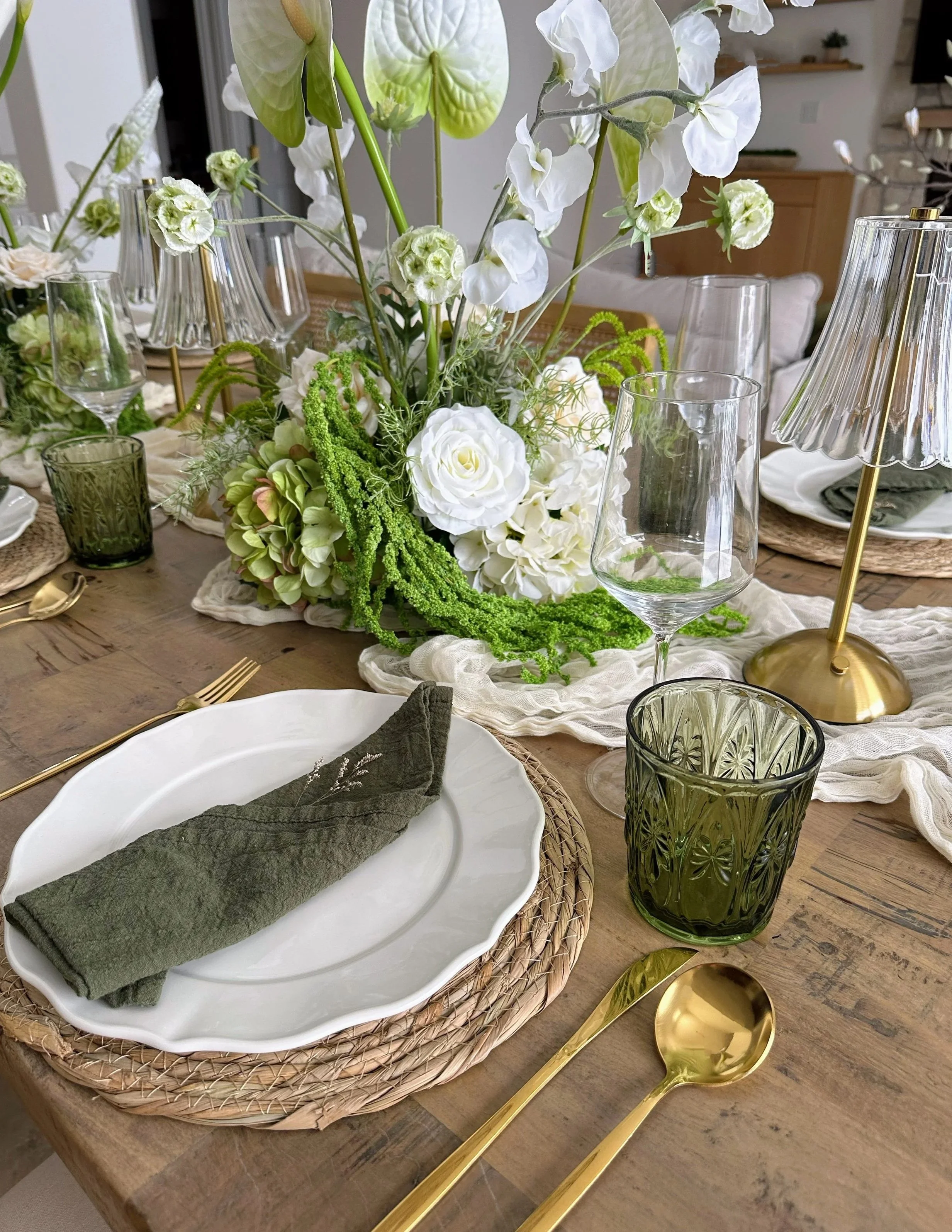Organic and earthy styled elegant dining table set with white tablecloth, floral centerpiece with green and white flowers, gold cutlery, green napkins, crystal glassware, and glass lamps.