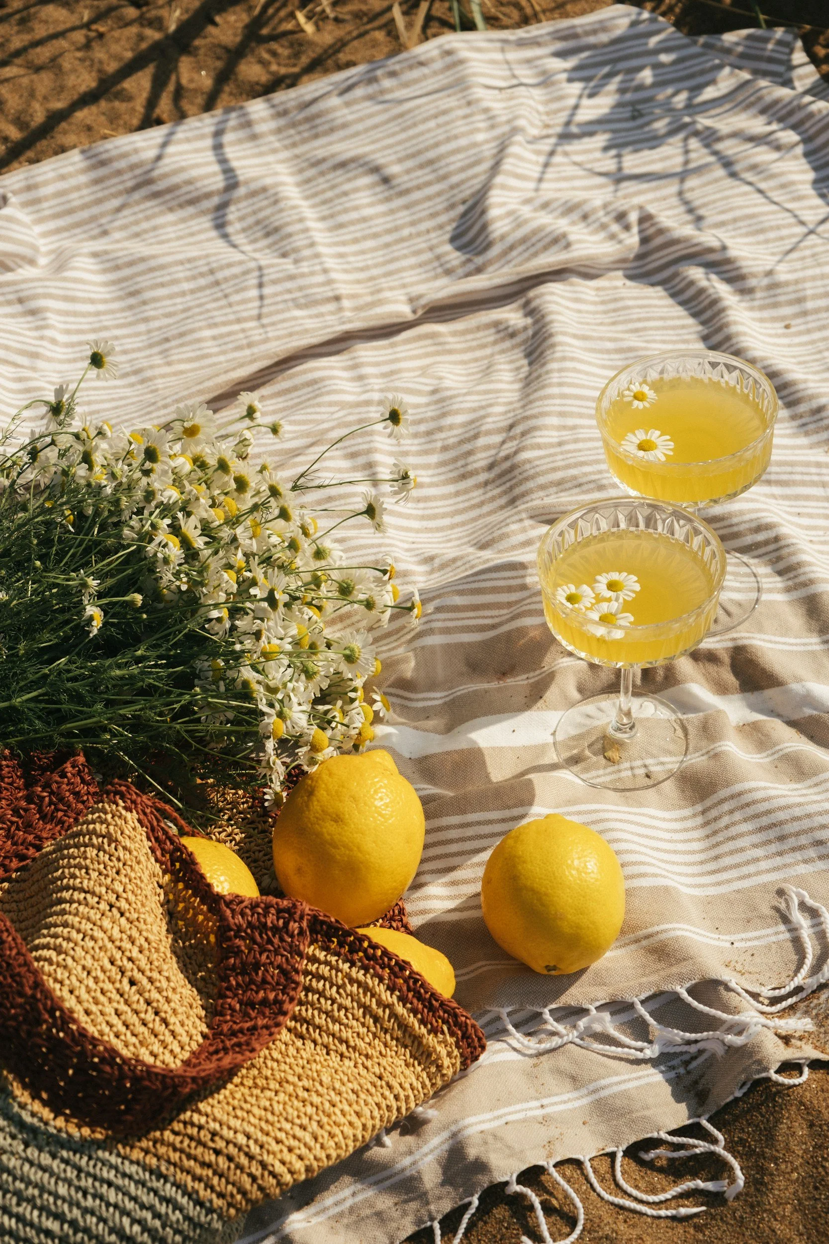 A picnic setup with two lemon cocktails in glass cups, a bouquet of daisies, fresh lemons, and a woven bag on a striped cloth on the ground outdoors.