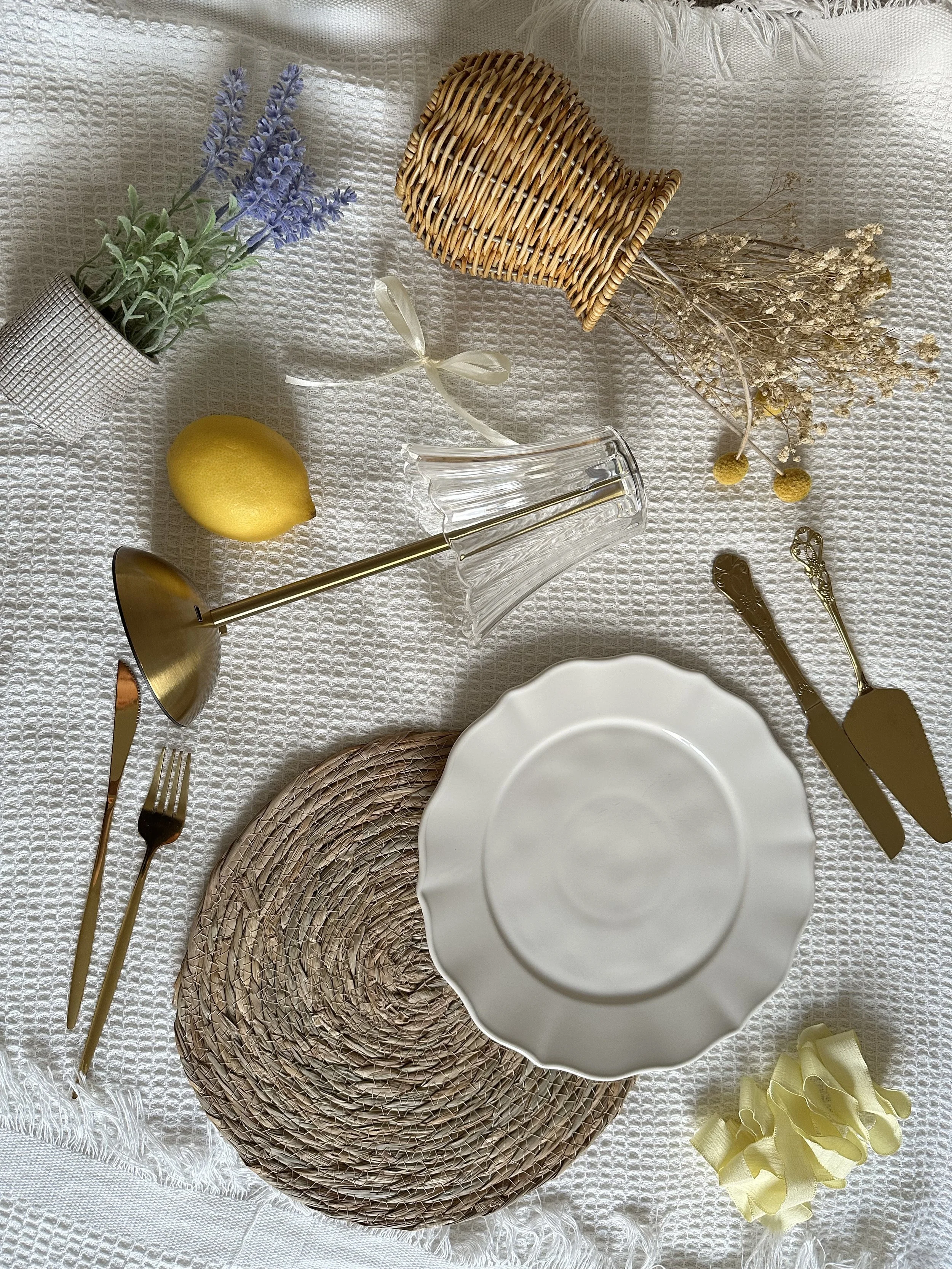 A table setting on a white textured tablecloth includes a white scalloped-edge plate, a woven placemat, gold flatware, a clear glass candlestick holder, a small woven basket with lavender flowers, a lemon, a bunch of dried flowers, and a yellow bow