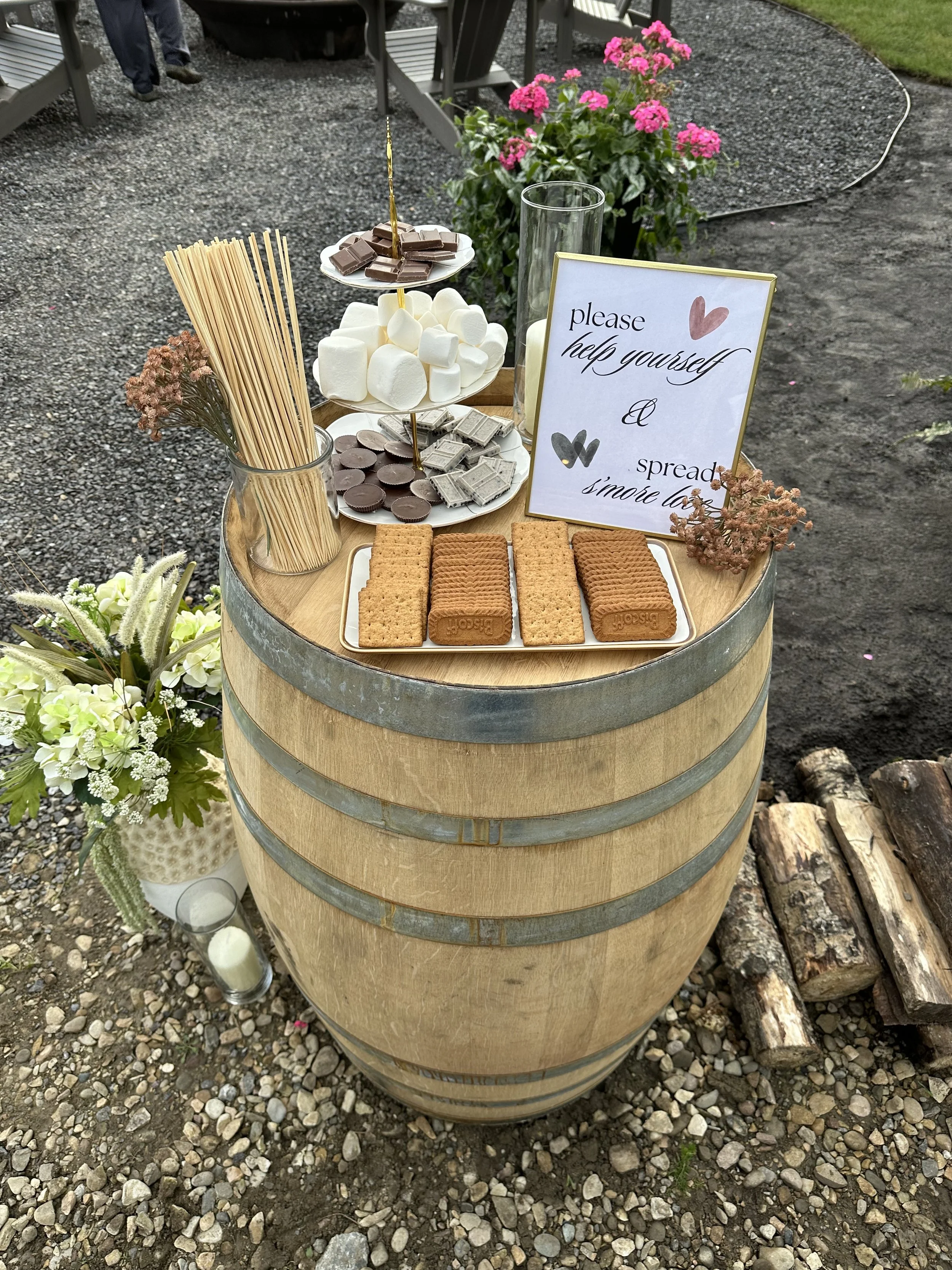 A wooden barrel used as a table with baked goods, marshmallows, chocolates, and graham crackers on plates. There are also flower arrangements, a sign with a message, and some candles on the ground nearby.