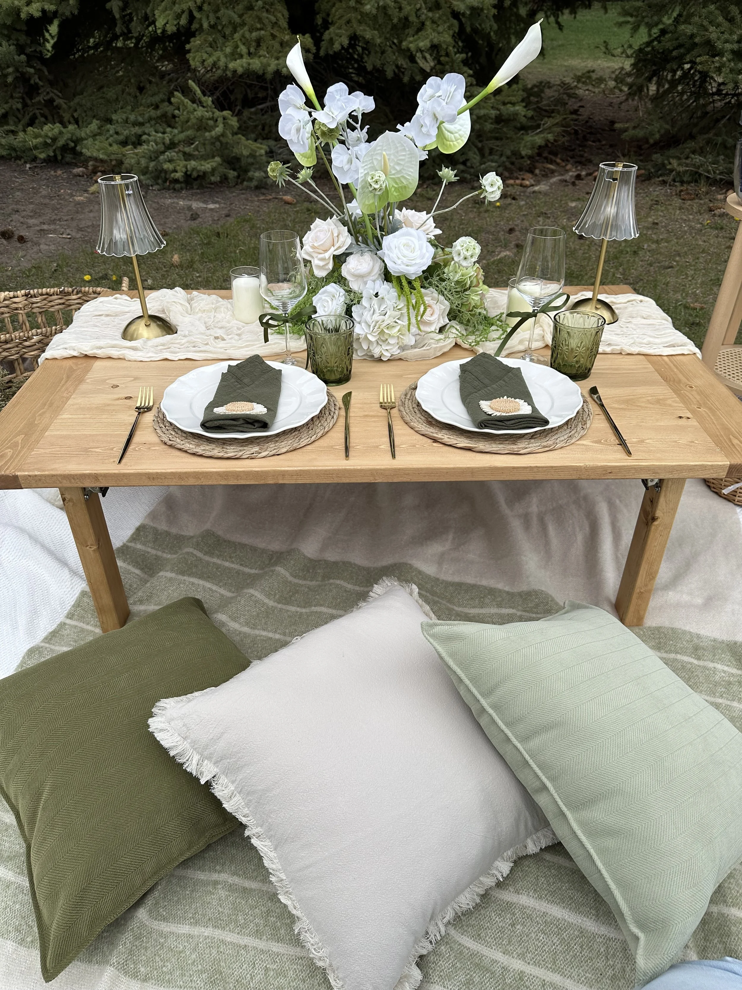 A wooden outdoor table set for a meal with white plates, silverware, green and white napkins, and a large floral centerpiece of white flowers. There are two small table lamps, candles, and glasses with water. Cushions are arranged on the ground for s