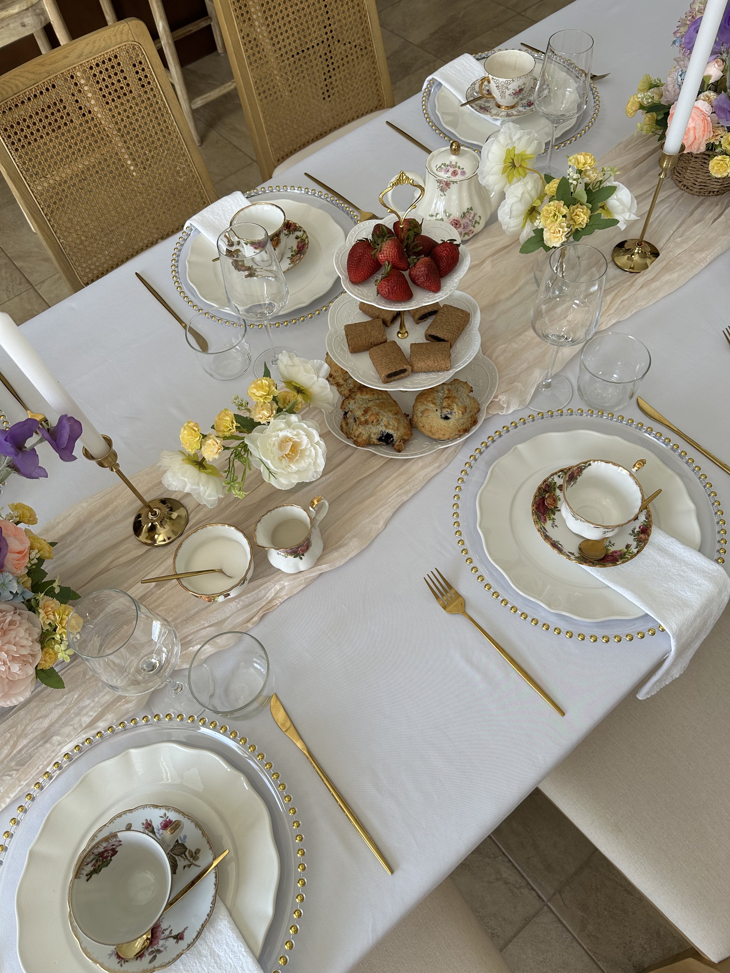 A beautifully set dining table with floral china plates, teacups, gold utensils, and a tiered stand with strawberries, cookies, and scones, decorated with flowers and candles for a special occasion.