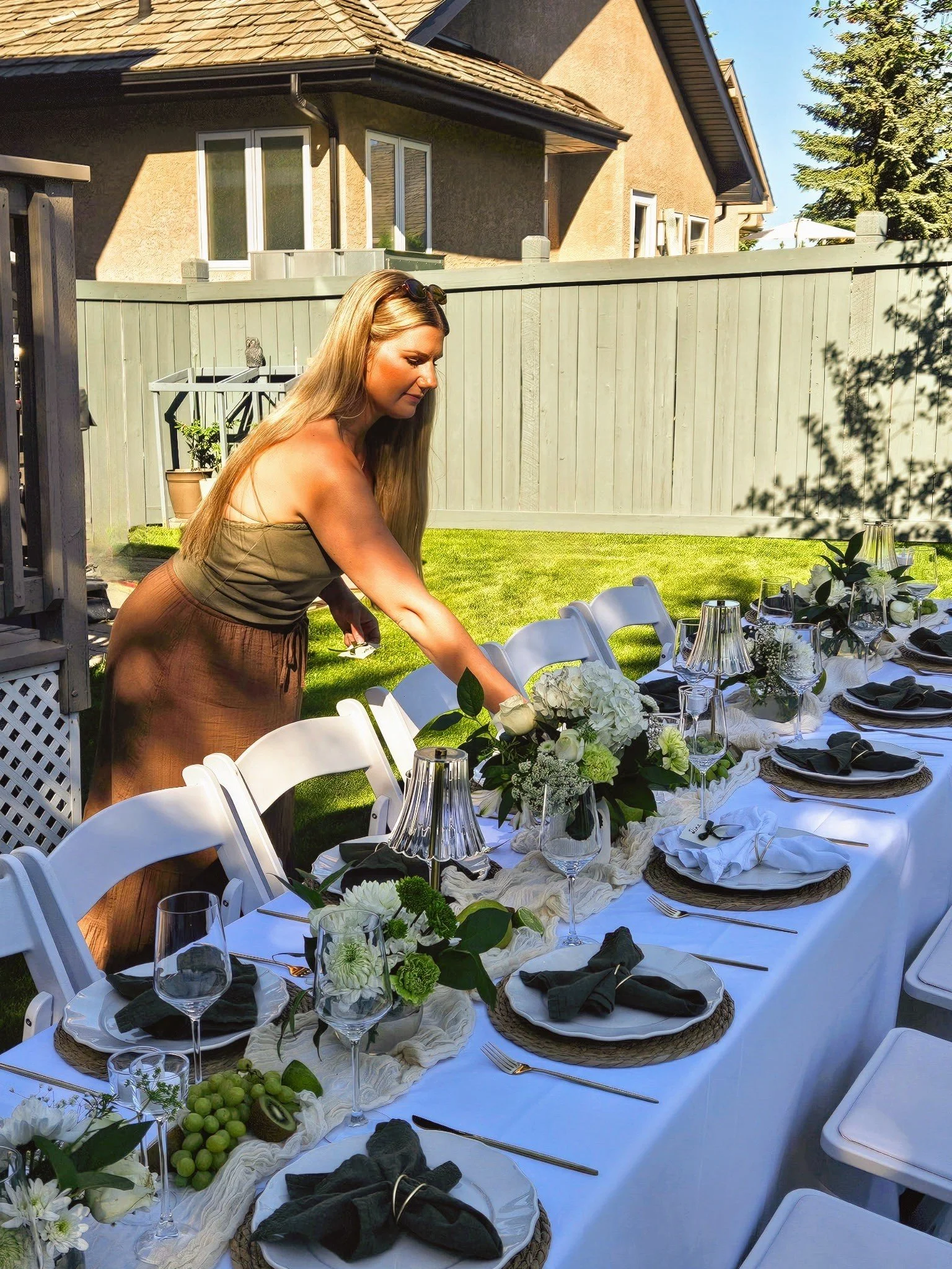 An event planner setting up a long dining table outdoors in a backyard, with white plates, olive napkins, wine glasses, and floral centerpieces, on a sunny day. Perfect for a garden dinner party event.