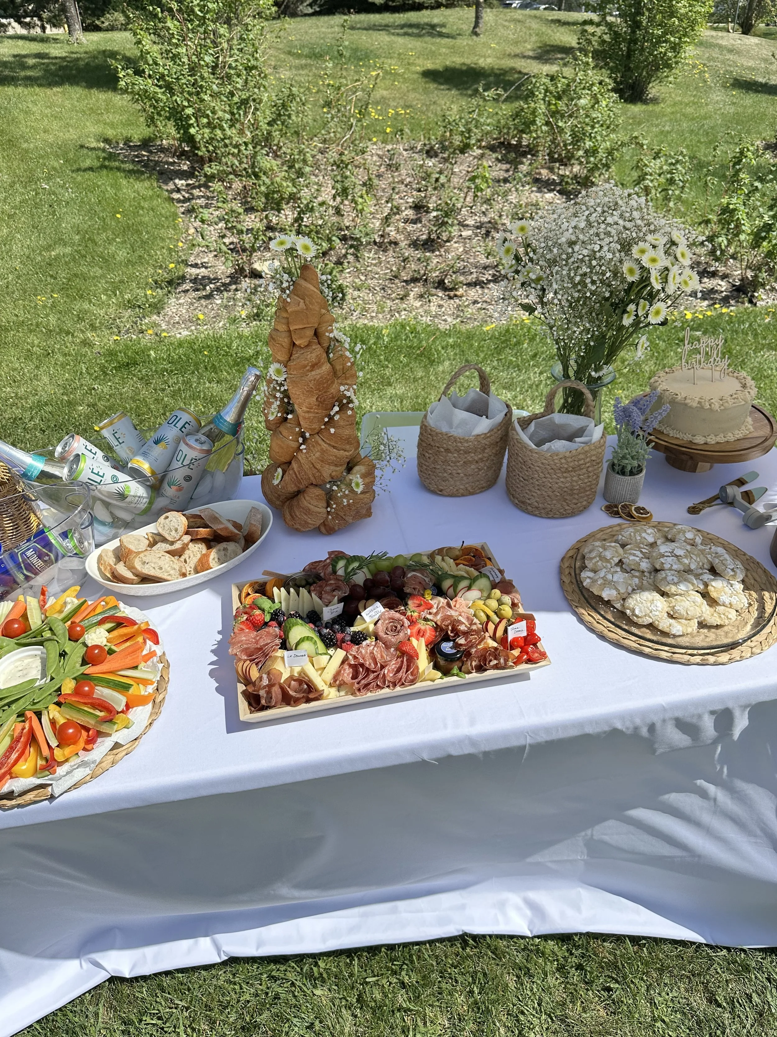 A table with a white tablecloth holding various foods including a vegetable platter, bread slices, a charcuterie board with meats, cheeses, grapes, and strawberries, a bouquet of white flowers, a cake with a "happy birthday" topper, and cans of spark