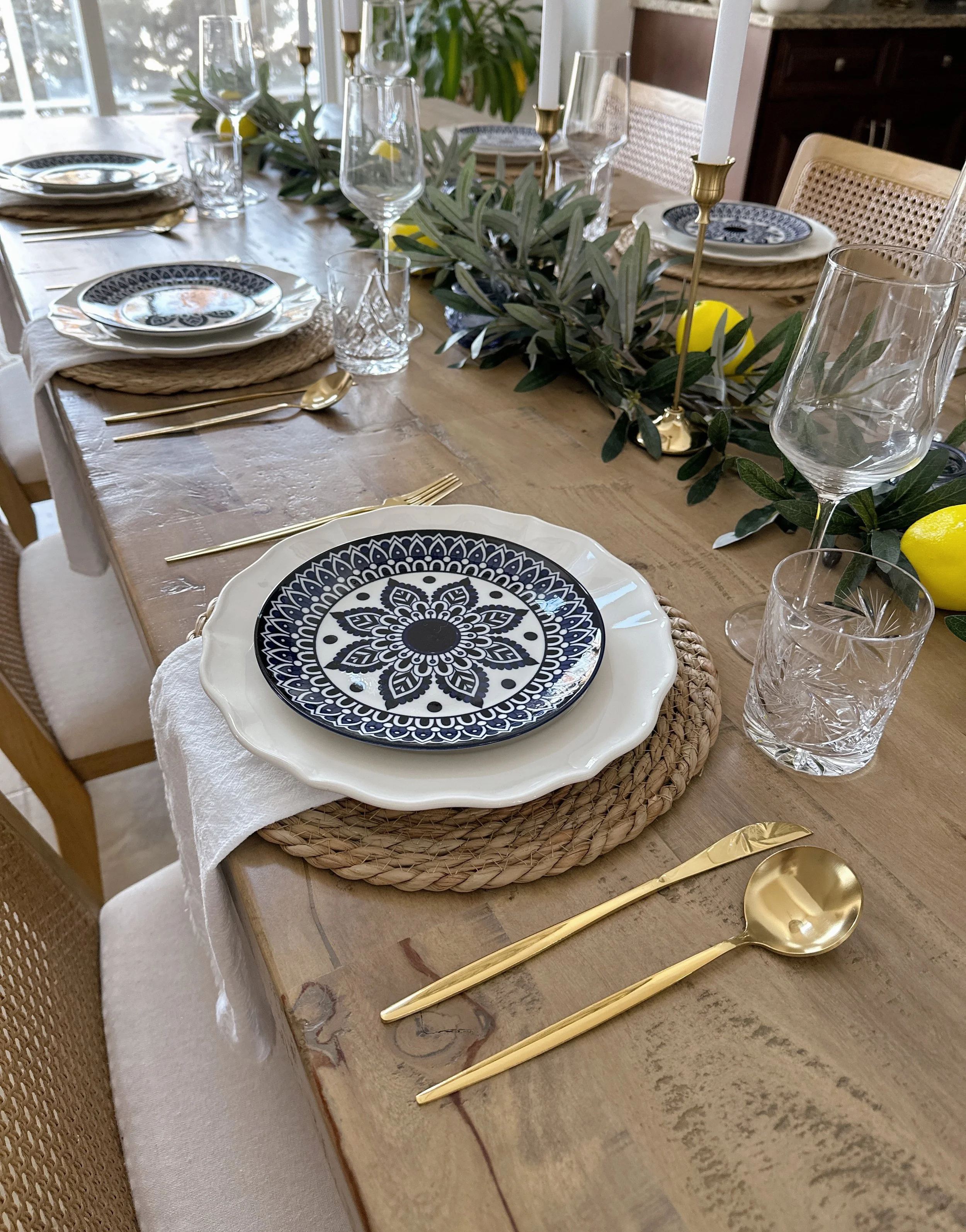 A decorated dining table with a natural wood surface, set with patterned blue and white plates, gold flatware, clear glassware, white cloth napkins, woven placemats, and a greenery and lemon centerpiece with tall white candles.