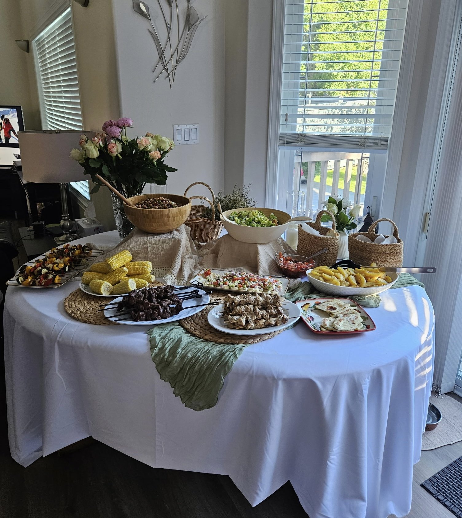 A round table covered with a white tablecloth holds various dishes and snacks, including skewered meats, corn on the cob, salads, and flatbreads, with greenery and flowers as decoration, set in a well-lit room with large windows and blinds.
