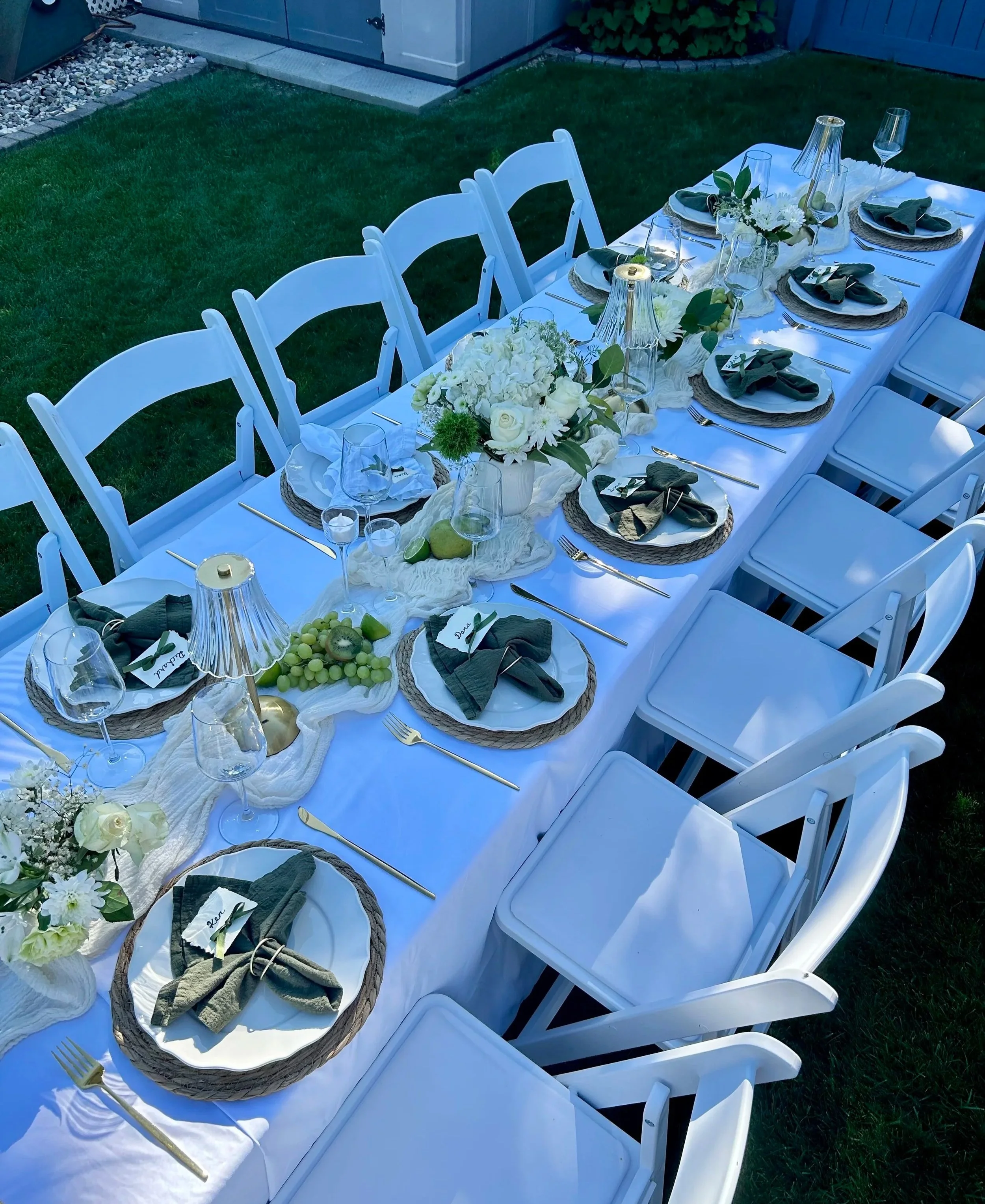 Outdoor dining table set for a birthday celebration, decorated with white and green flowers, green leaves, decorated with green fruit pears grapes and lime wedges, and fancy tableware, surrounded by white chairs on grass.
