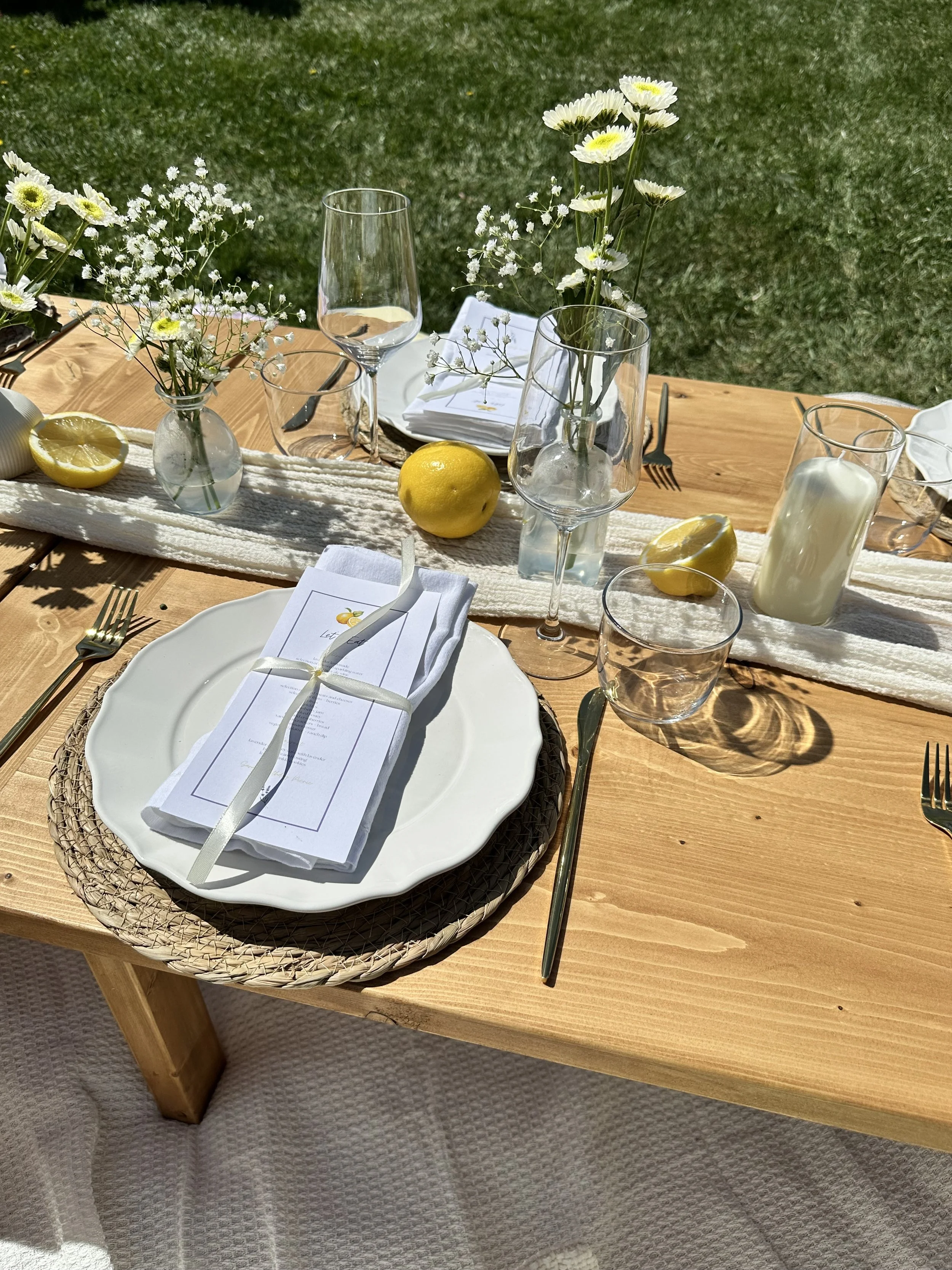 A wooden table set for a meal outdoors with white flowers in glass vases, lemons, a candle, and place settings with plates, utensils, napkins, and menus.