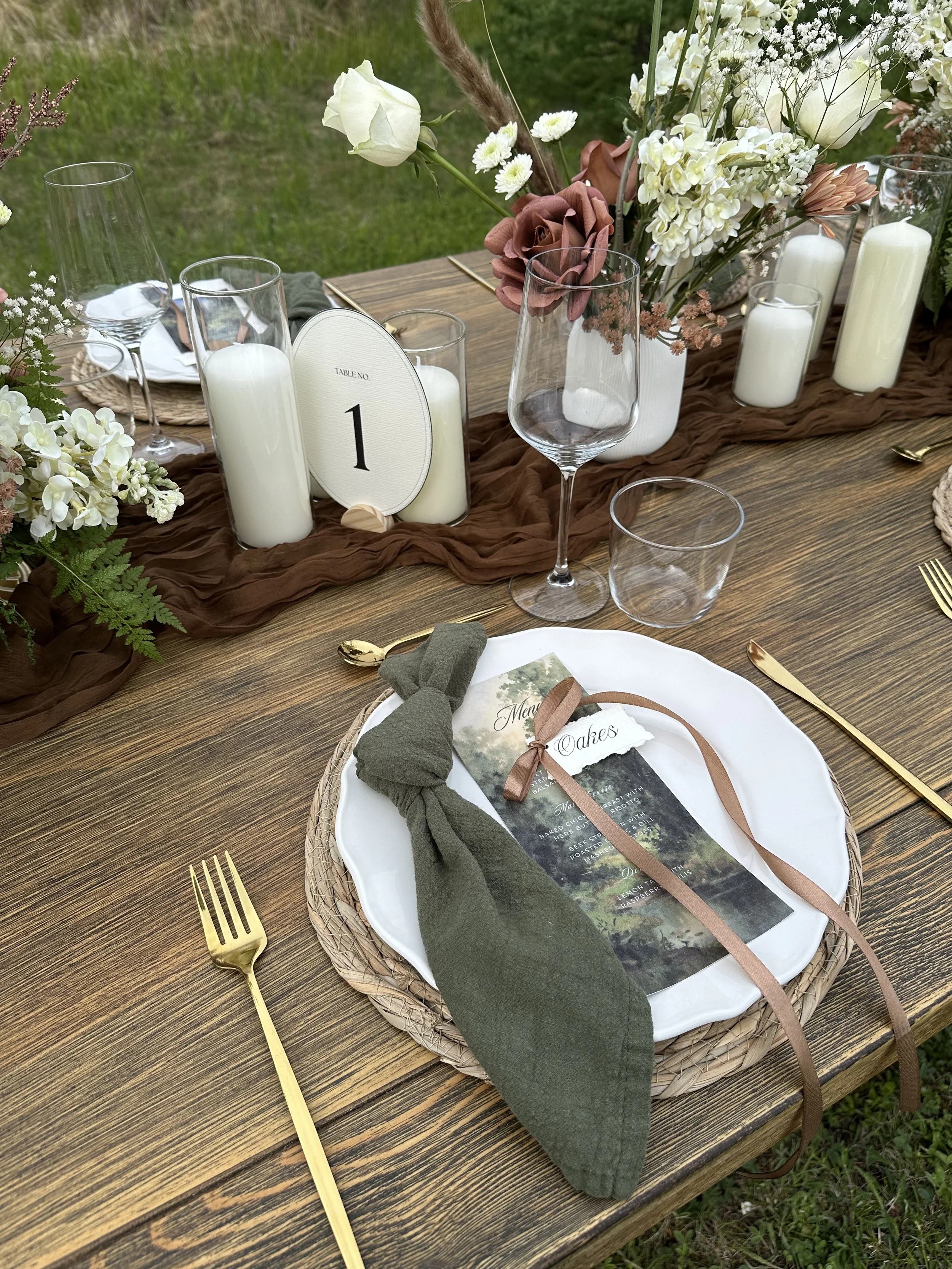 A rustic outdoor table decorated with a brown table runner, a floral centerpiece with white and blush flowers, glass candles, and a place setting with a white plate, gold utensils, a menu, and a forest-themed napkin bundle tied with Ribbon.