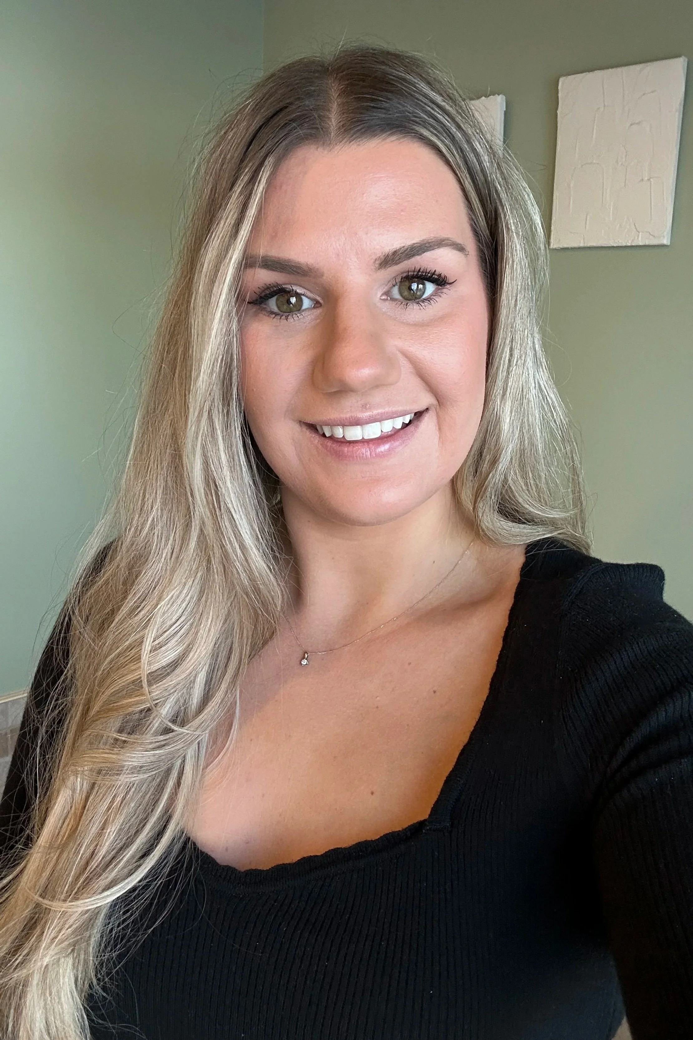 A smiling woman with long blonde hair, wearing a black top and a delicate necklace, standing indoors with a green wall and white artwork behind her.