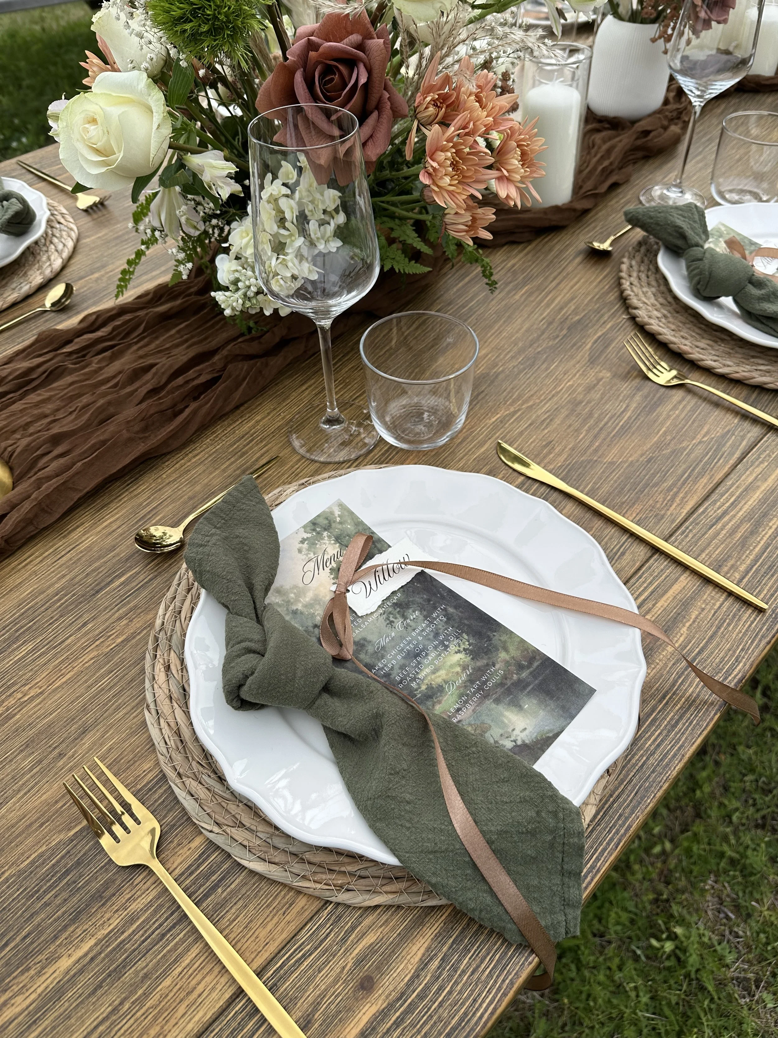 A table set for a formal outdoor event with a floral centerpiece, gold flatware, white plates, a menu card, green napkin, and a rustic brown table runner. The centerpiece includes roses, chrysanthemums, and other flowers in shades of cream, peach, an