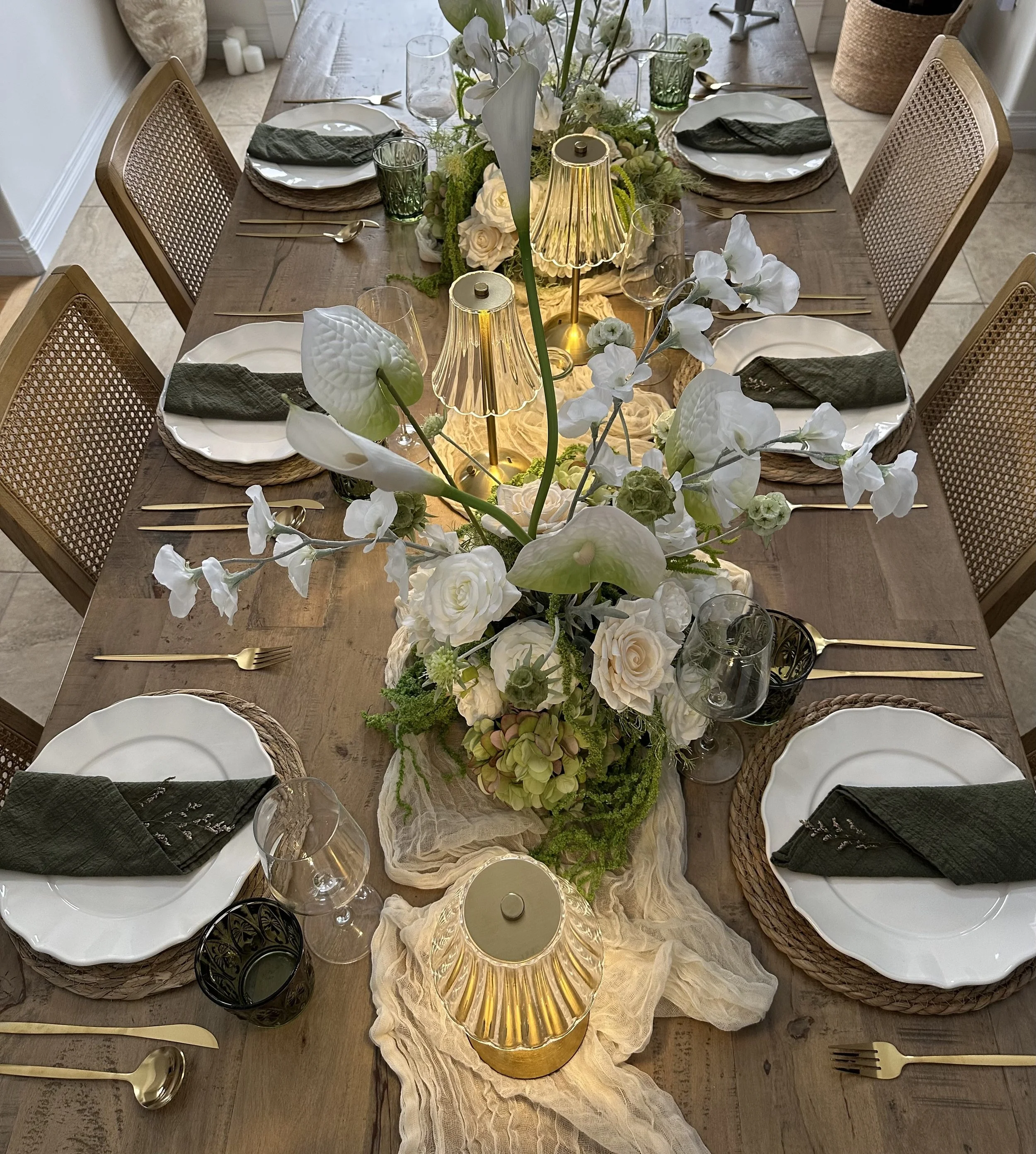 A rectangular dining table set for six people with white plates, dark napkins, gold utensils, and various drinking glasses. The table features a central floral arrangement with white roses, orchids, and greenery, and has three vintage-style lamps wit