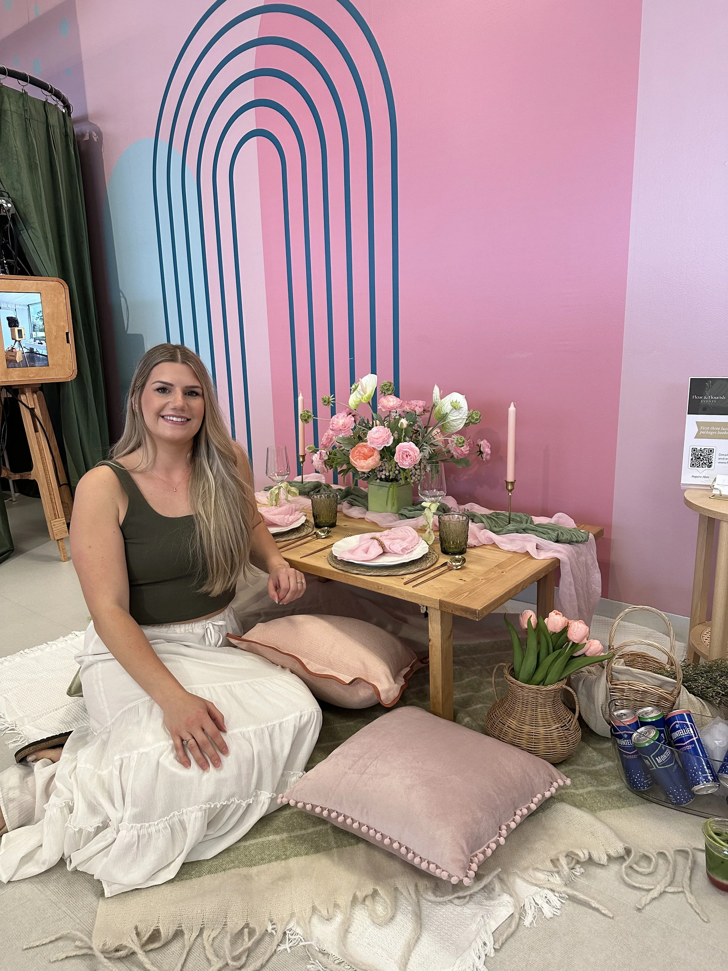 A woman with long blonde hair is sitting on a beige and white rug, smiling at the camera. She is in front of a decorated table with pink and white flowers, candles, and glasses. The background features a pink wall with a blue rainbow mural, green cur