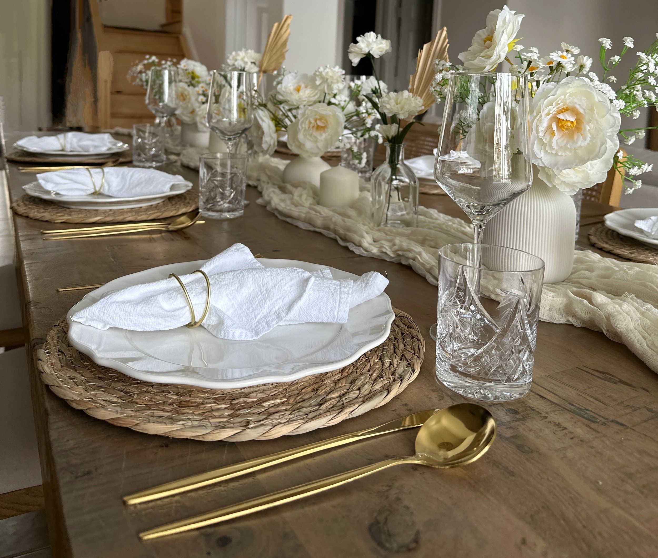 Elegant dining table setup with white plates, gold cutlery, glassware, white cloth napkins, woven placemats, and floral centerpieces with white flowers in vases.