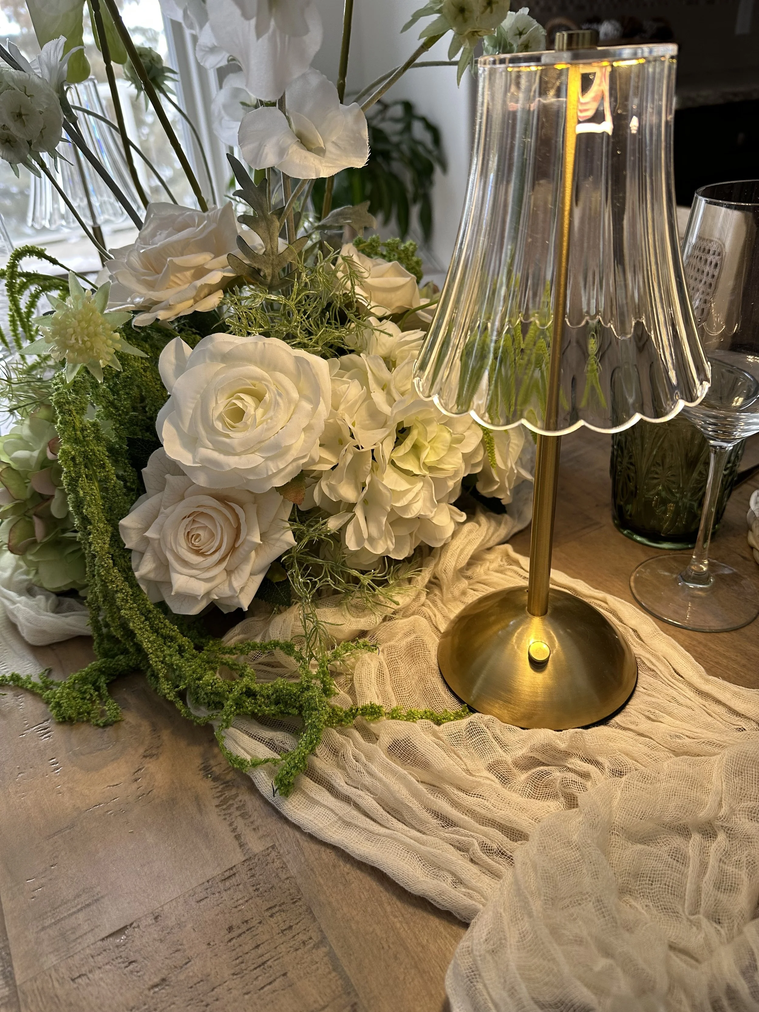A floral arrangement with white roses, ranunculus, and greenery on a table with a lace tablecloth. A gold lamp with a scalloped glass shade is also on the table, along with glasses and a dark green bottle in the background.