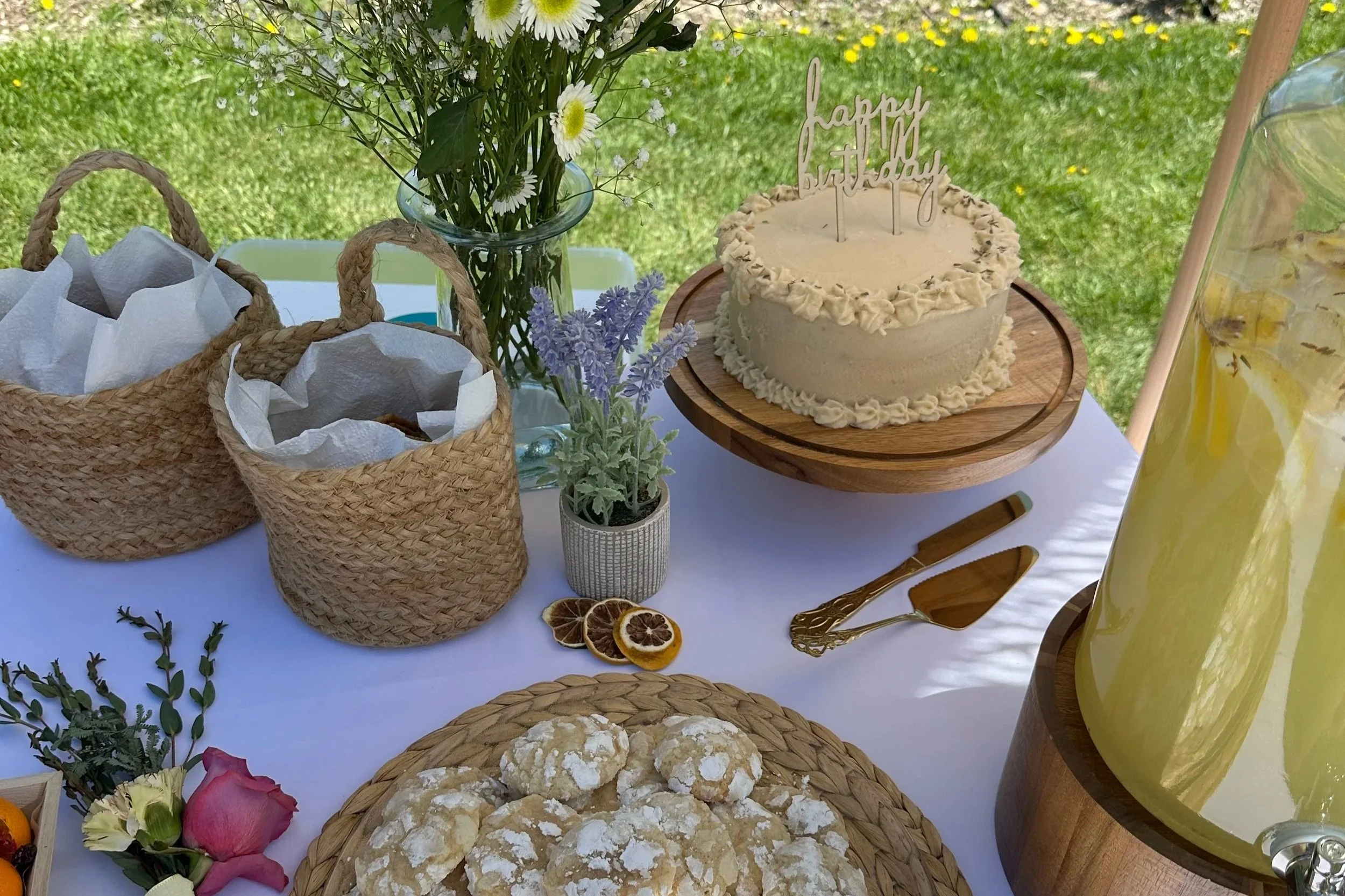 A food buffet table decorated for a birthday celebration with flowers, a birthday cake, desserts, and lemonade.