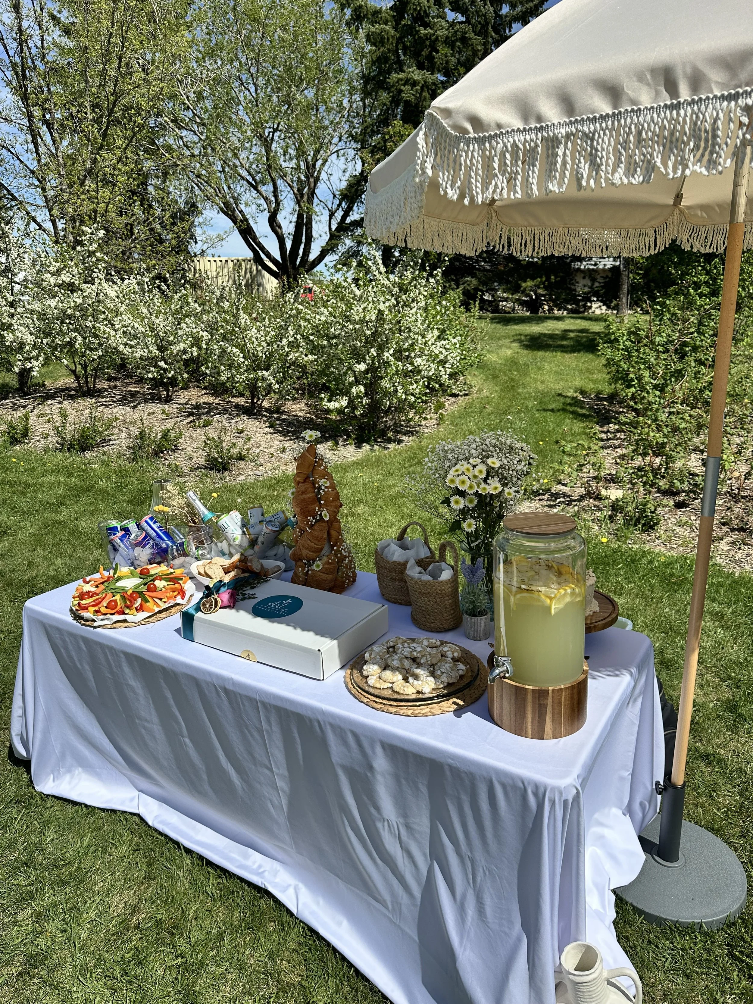 A table with snacks and drinks under a white umbrella in a garden with trees and flowering bushes.