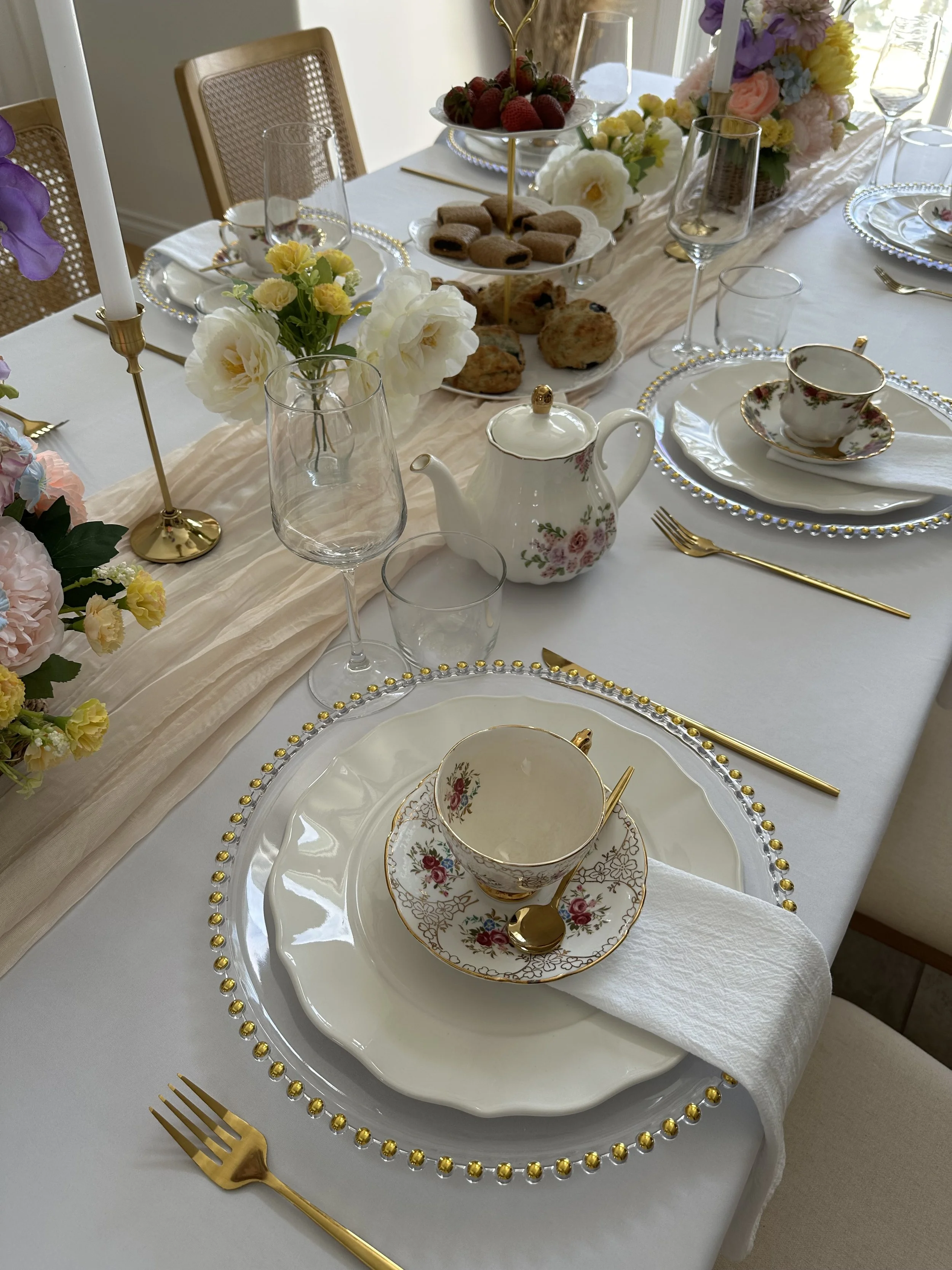 A formal dining table set with white and gold dinnerware, gold utensils, glassware, and floral centerpieces, with a buffet of cookies, strawberries, and scones.