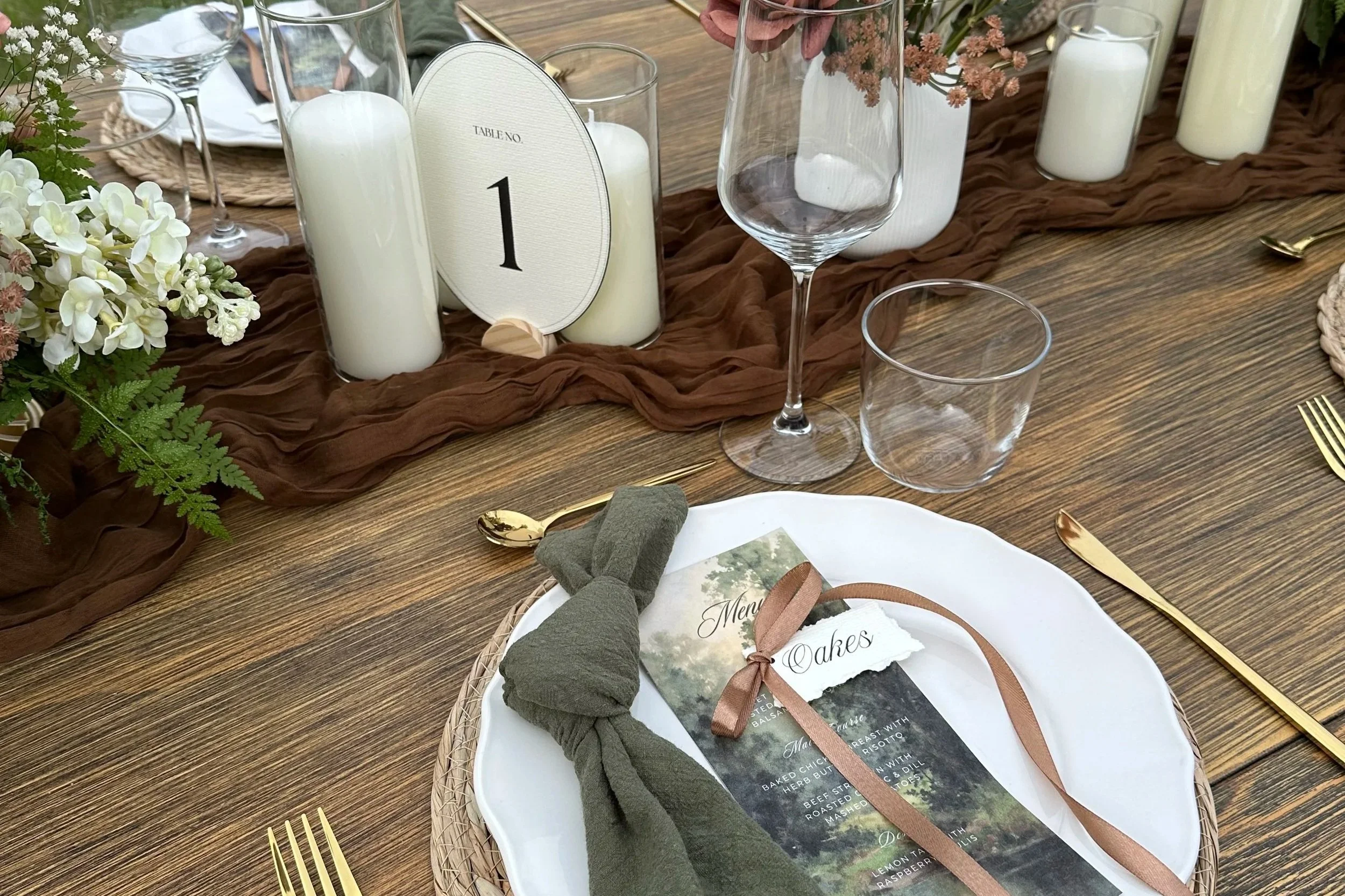A decorated outdoor woodsy wedding reception table setup with a moody color palette of olive, brown, and ivory. Decorated with a brown table runner, candles, floral arrangements, glasses, gold utensils, and stationery menu and place card with ribbon.