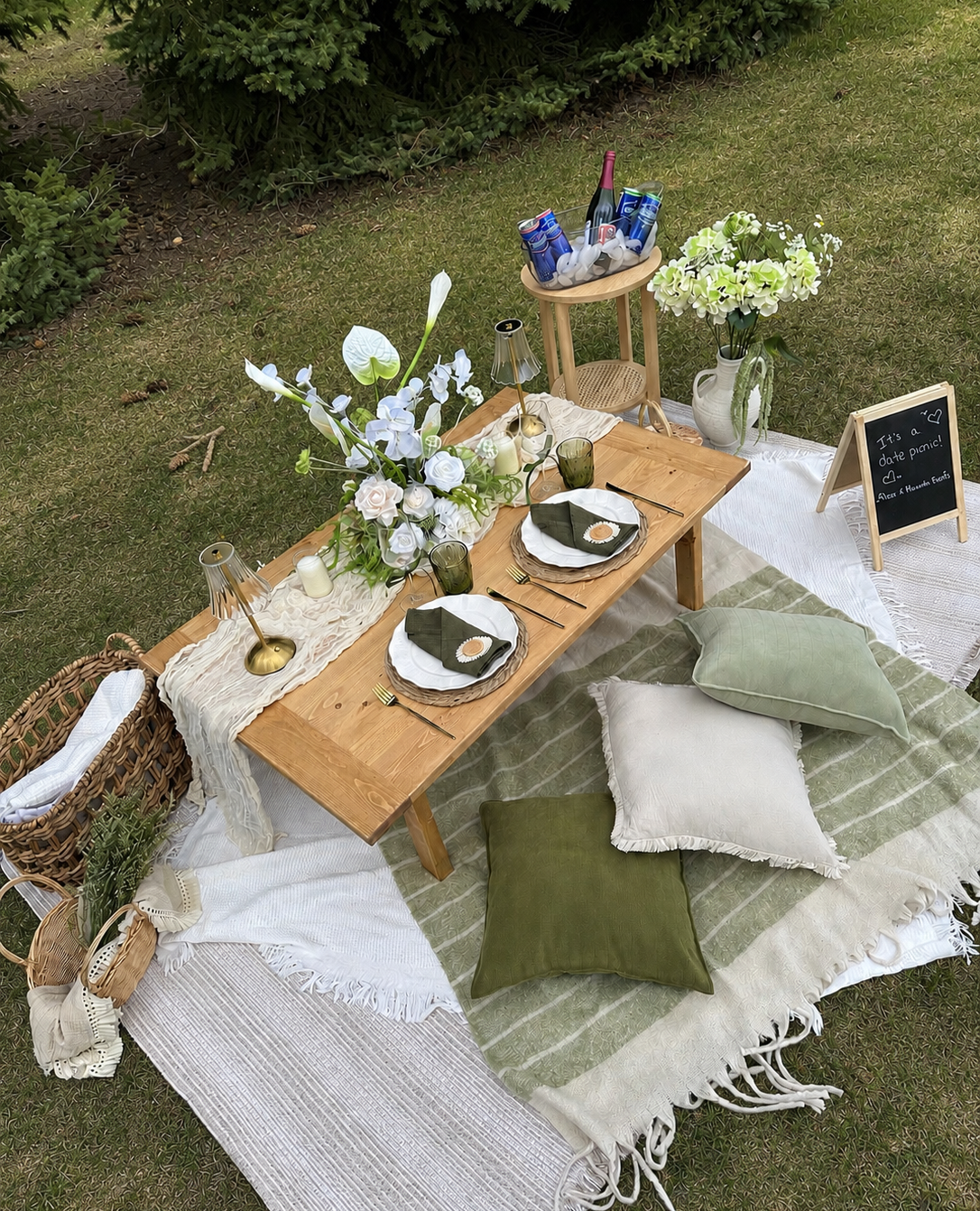 An outdoor luxury picnic set up with a natural, organic theme featuring greens and whites. Includes green drinking glasses, wine glasses, candles, and brass lamps .