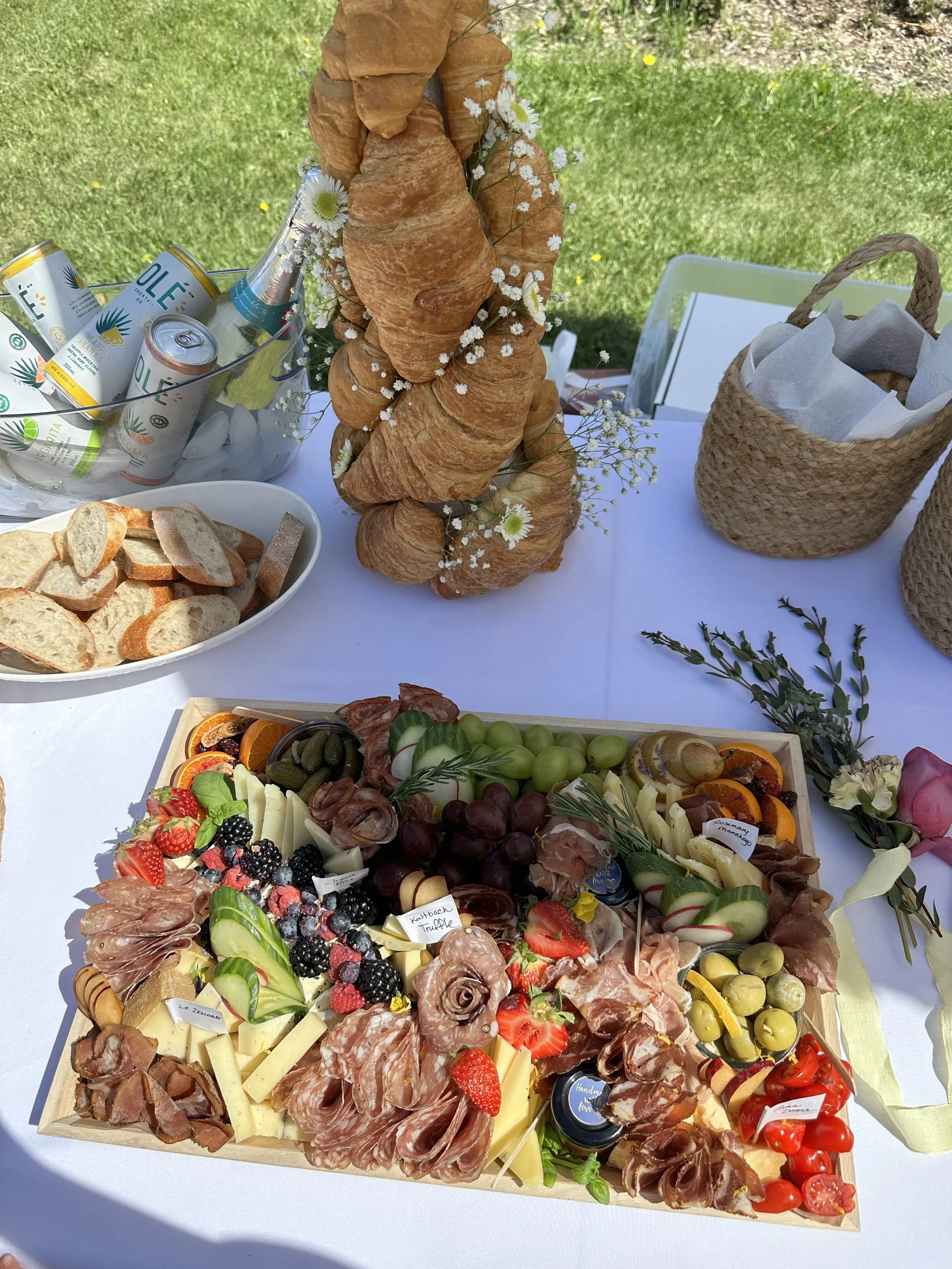 A charcuterie board with cheese, meats, fresh fruit, and berries, along with a basket of wrapped items and herbs. In the background, there is a display of croissants, canned drinks, and a floral decoration.